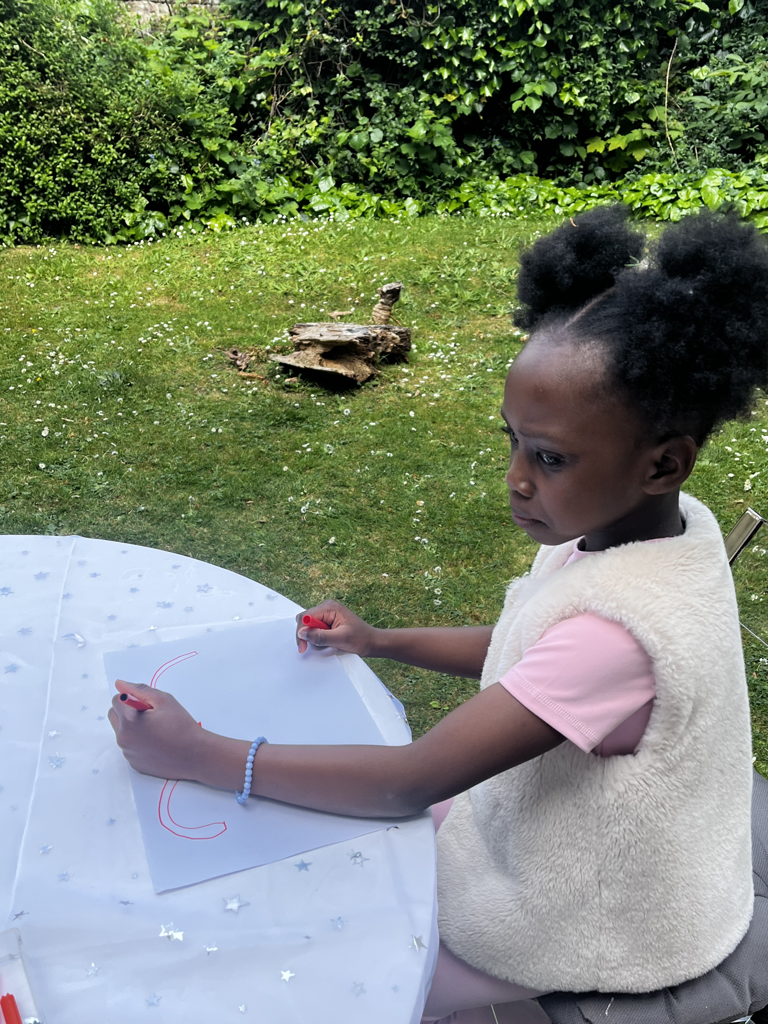 A young girl with curly hair sits at a table outside, drawing on a piece of paper with a red marker. She is wearing a pink shirt with a cream-colored vest and a bracelet. In the background, there is a lawn with small white flowers, a fallen piece of wood, and dense green bushes.