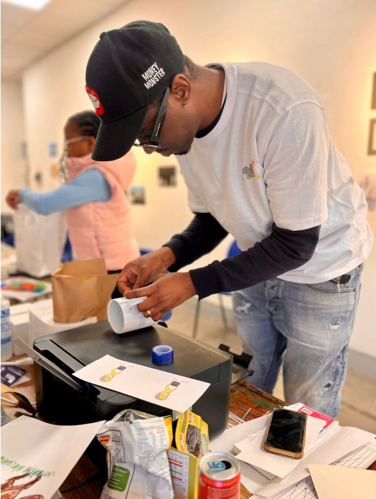 A man wearing glasses, a black cap, a white t-shirt, and jeans is working on a project at a table, with a woman behind him, in an indoor setting. The table is cluttered with various items, including paper, tape, a mug, and snacks.