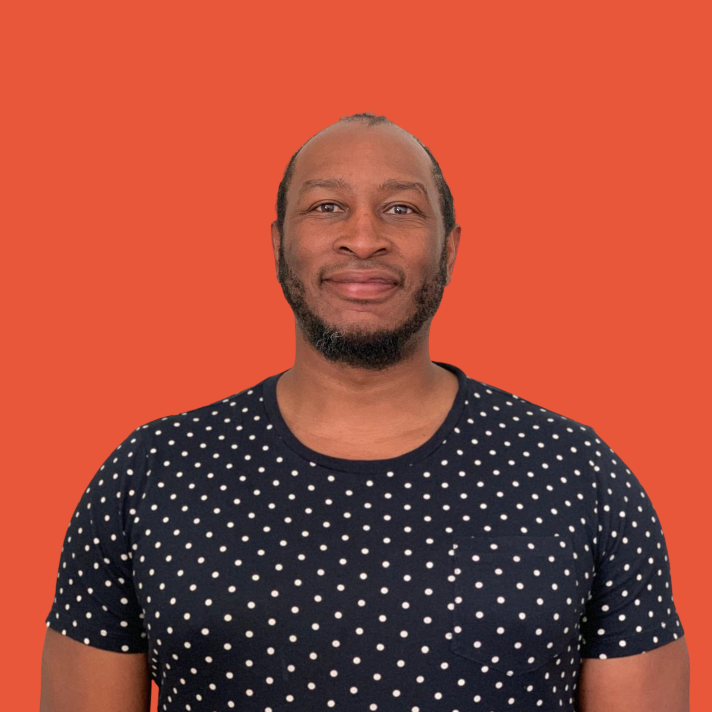 A man with a beard and short hair smiling, wearing a navy blue t-shirt with white polka dots, against an orange background.