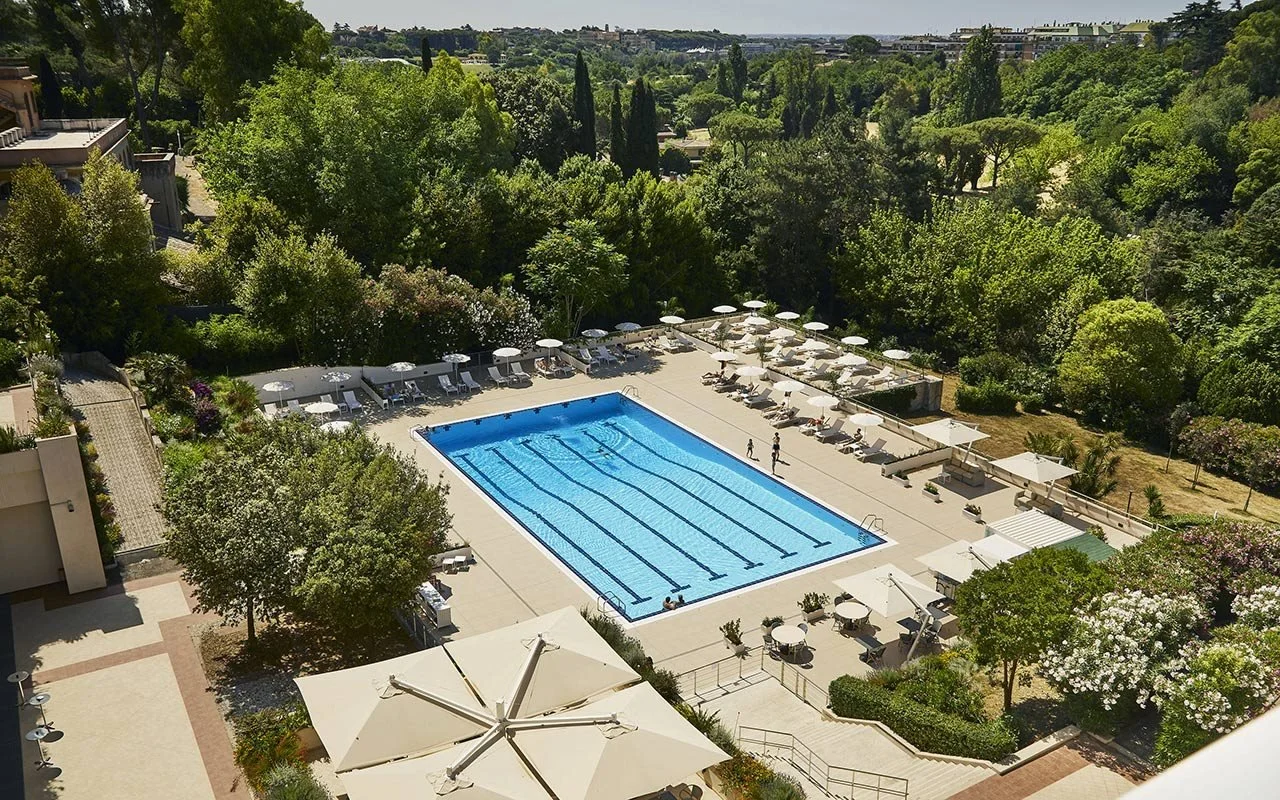 Outdoor 25-meter pool with loungers and umbrellas surrounded by trees at Hotel Villa Pamphili Roma