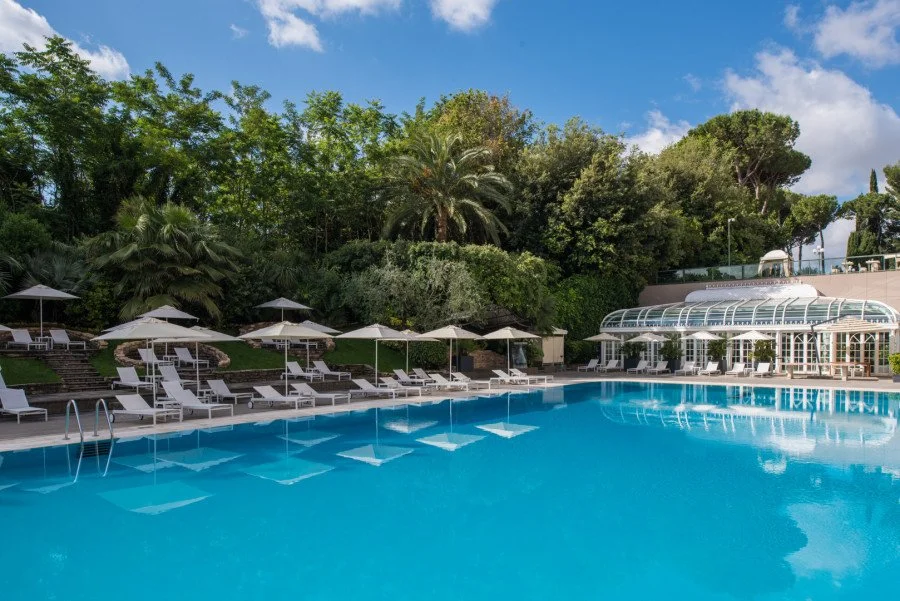 Large outdoor pool with white loungers and umbrellas surrounded by gardens at Rome Cavalieri, Monte Mario
