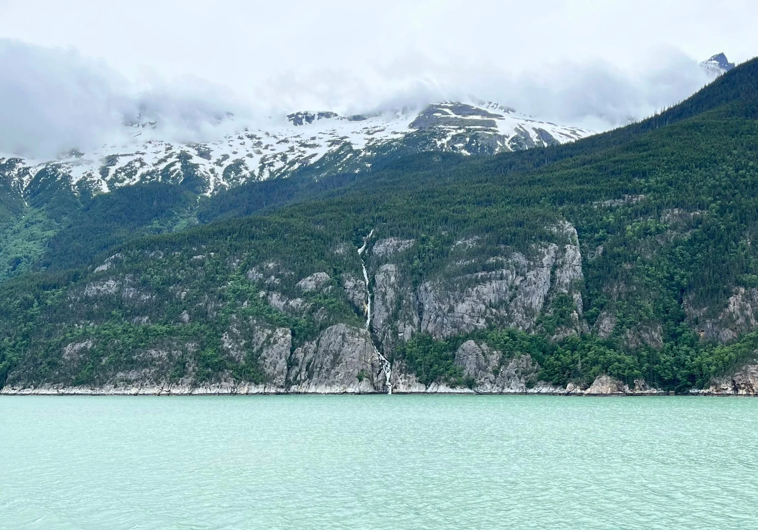 Scenic landscape with a lake in the foreground, a forested mountainside with a waterfall, and snow-capped peaks in the background under an overcast sky. Alaska has got it all. Sun, Snow, Mountains, water, clouds, and endless space. Enjoy water.