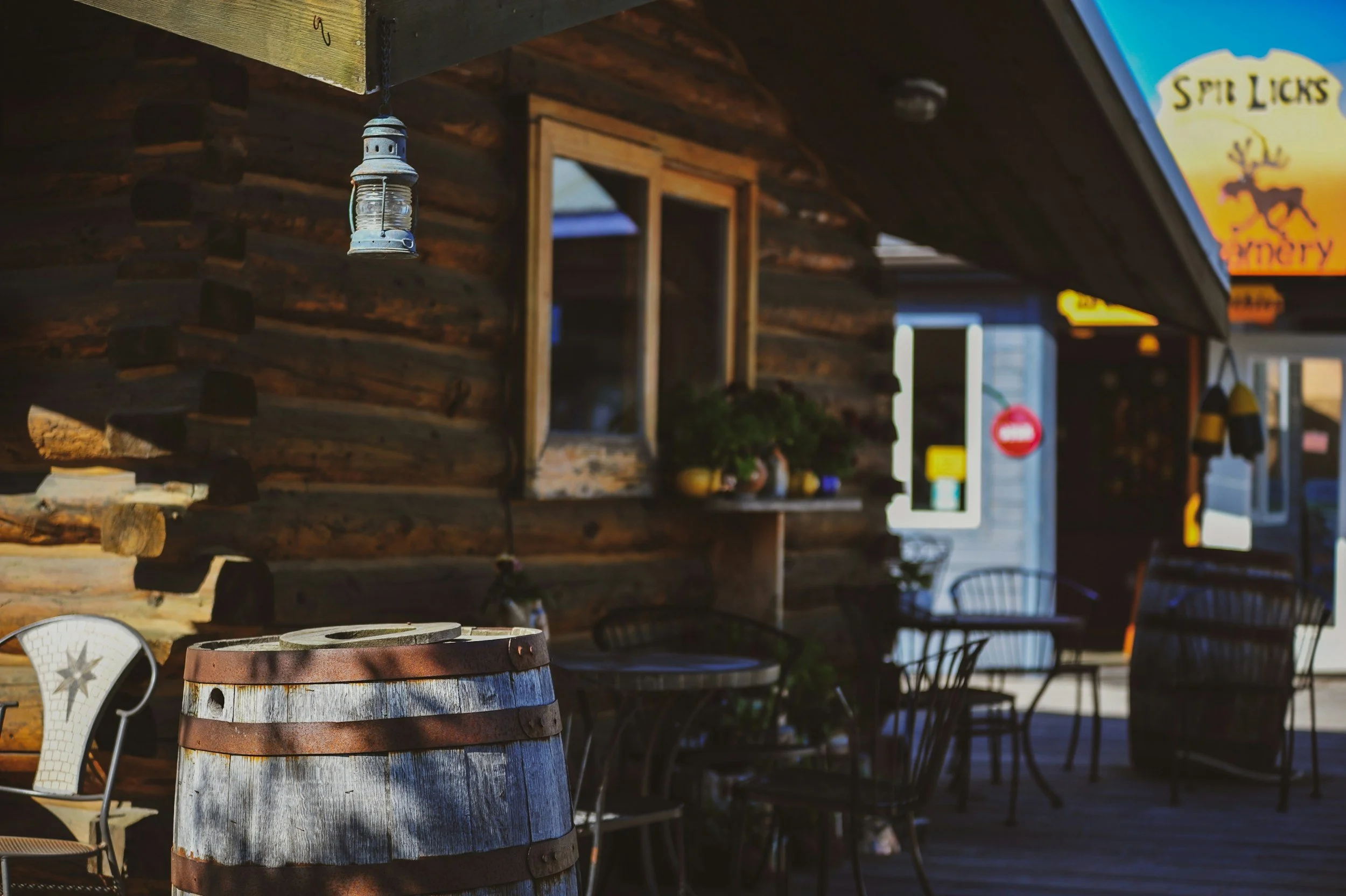 Outdoor seating area of a rustic restaurant or cafe, featuring a wooden barrel, metal chairs, a lantern hanging from the ceiling, and a log cabin-style building with a window and decorative plants.