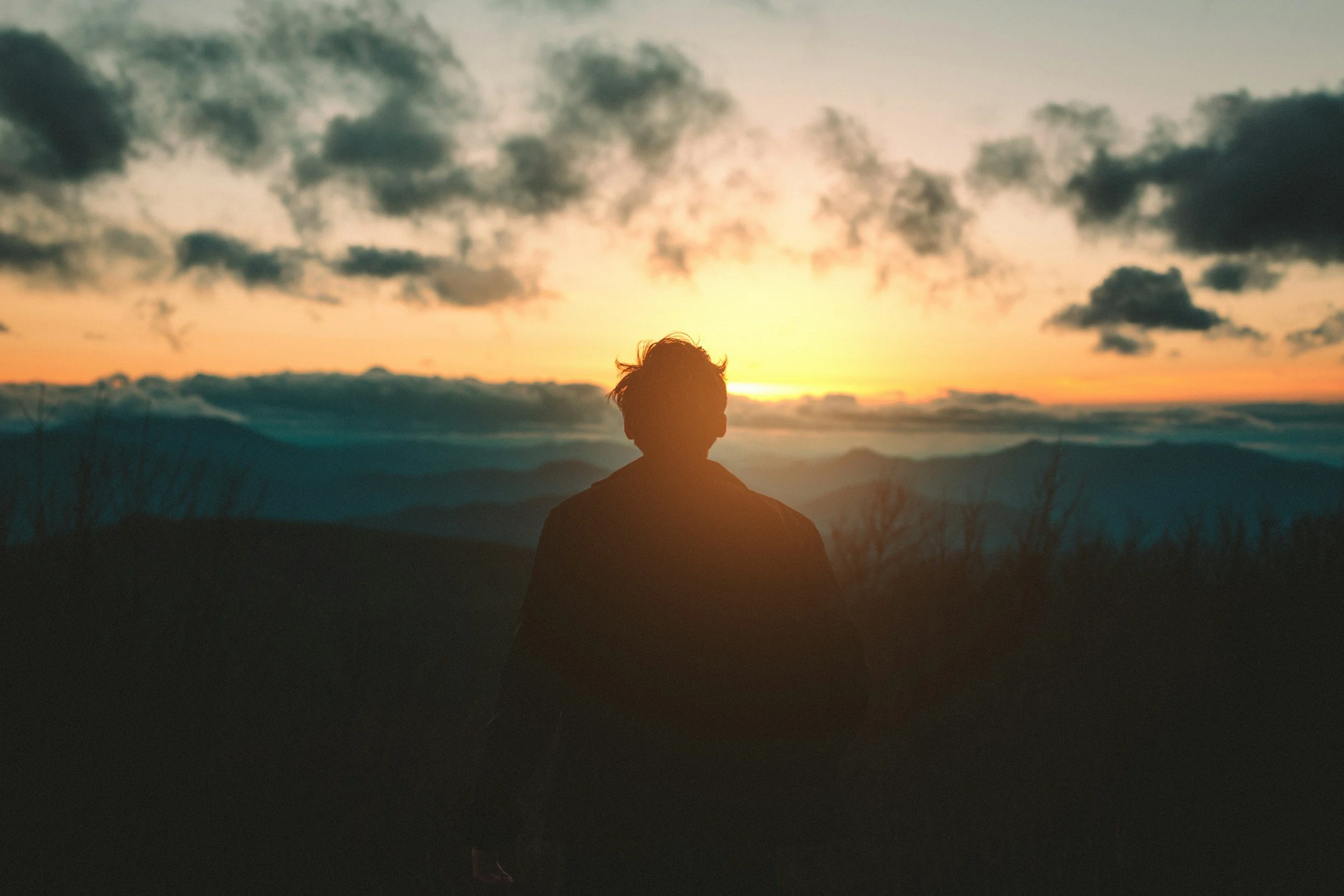 Silhouette of a person standing outdoors during sunset, overlooking mountains and clouds in the sky.