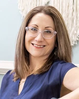 A woman with shoulder-length brown hair wearing clear glasses and a blue top, smiling at the camera in a bright room with a beige wall and decorative wall hanging.