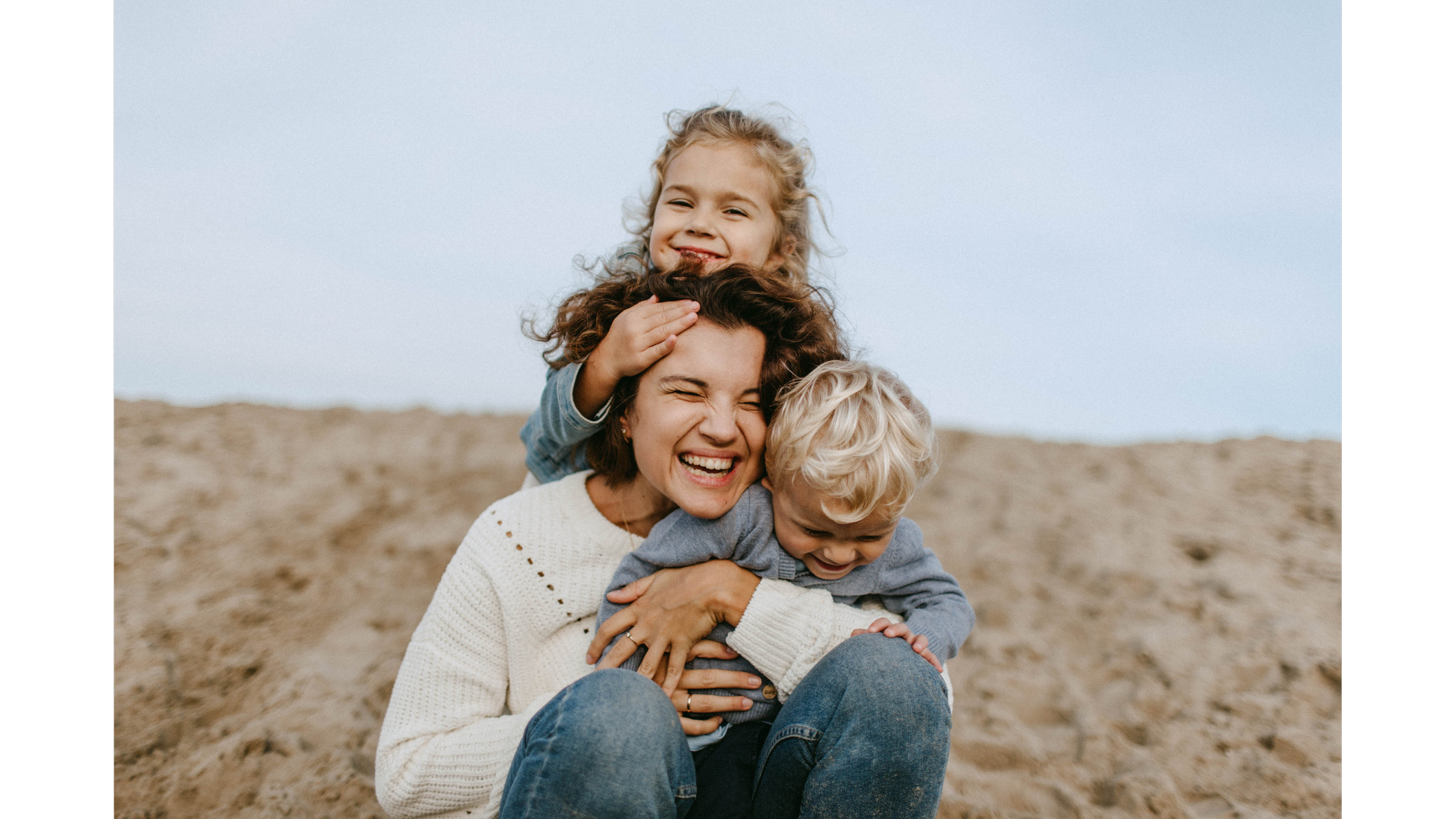 A woman with short brown hair smiling and laughing while holding two children, one child with curly blonde hair and another with straight blonde hair, on a sandy beach during daytime.