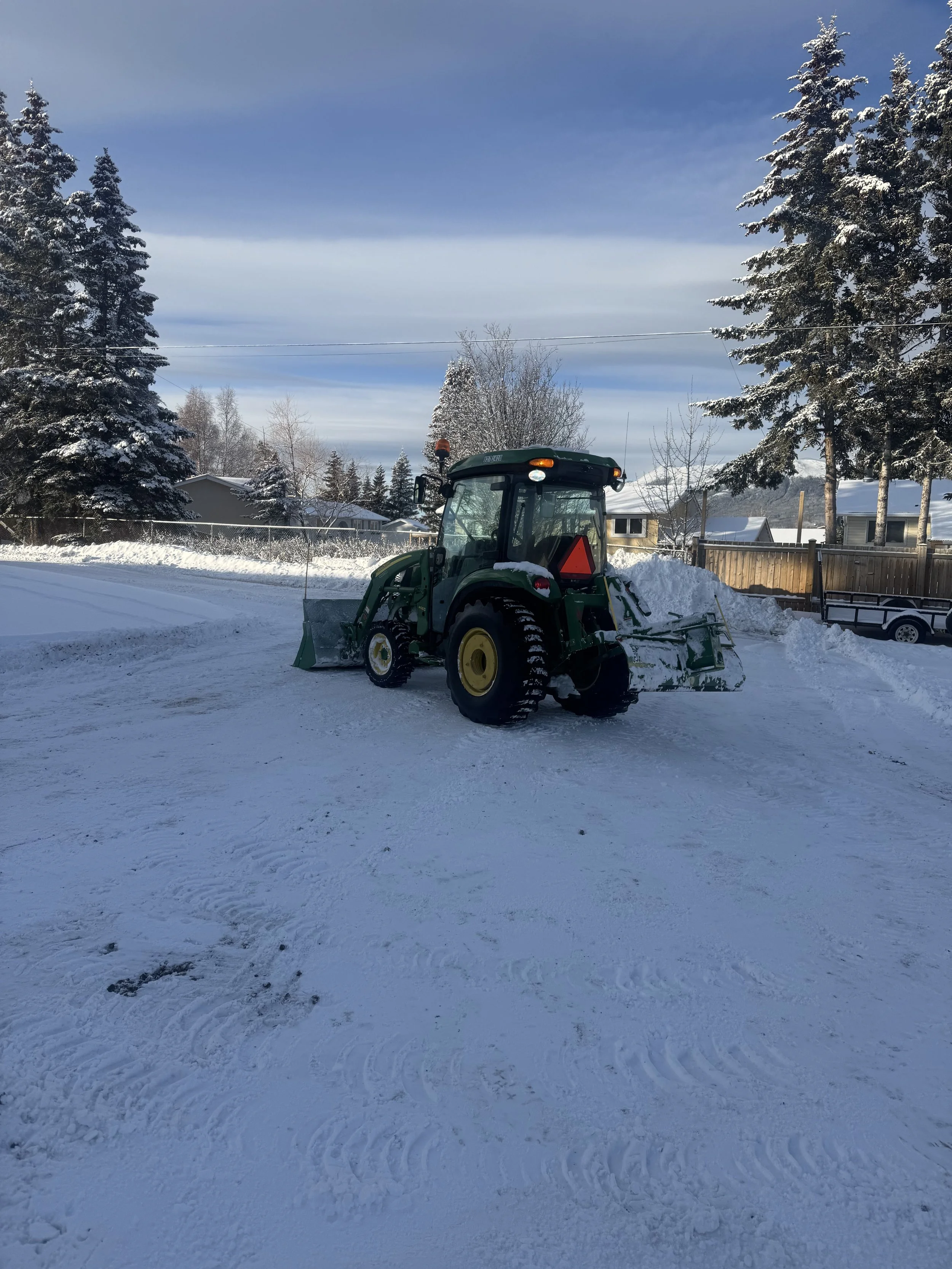 A green tractor with snow on the front blade is clearing a snow-covered driveway in a residential neighborhood with snow-covered trees and houses under a partly cloudy sky.