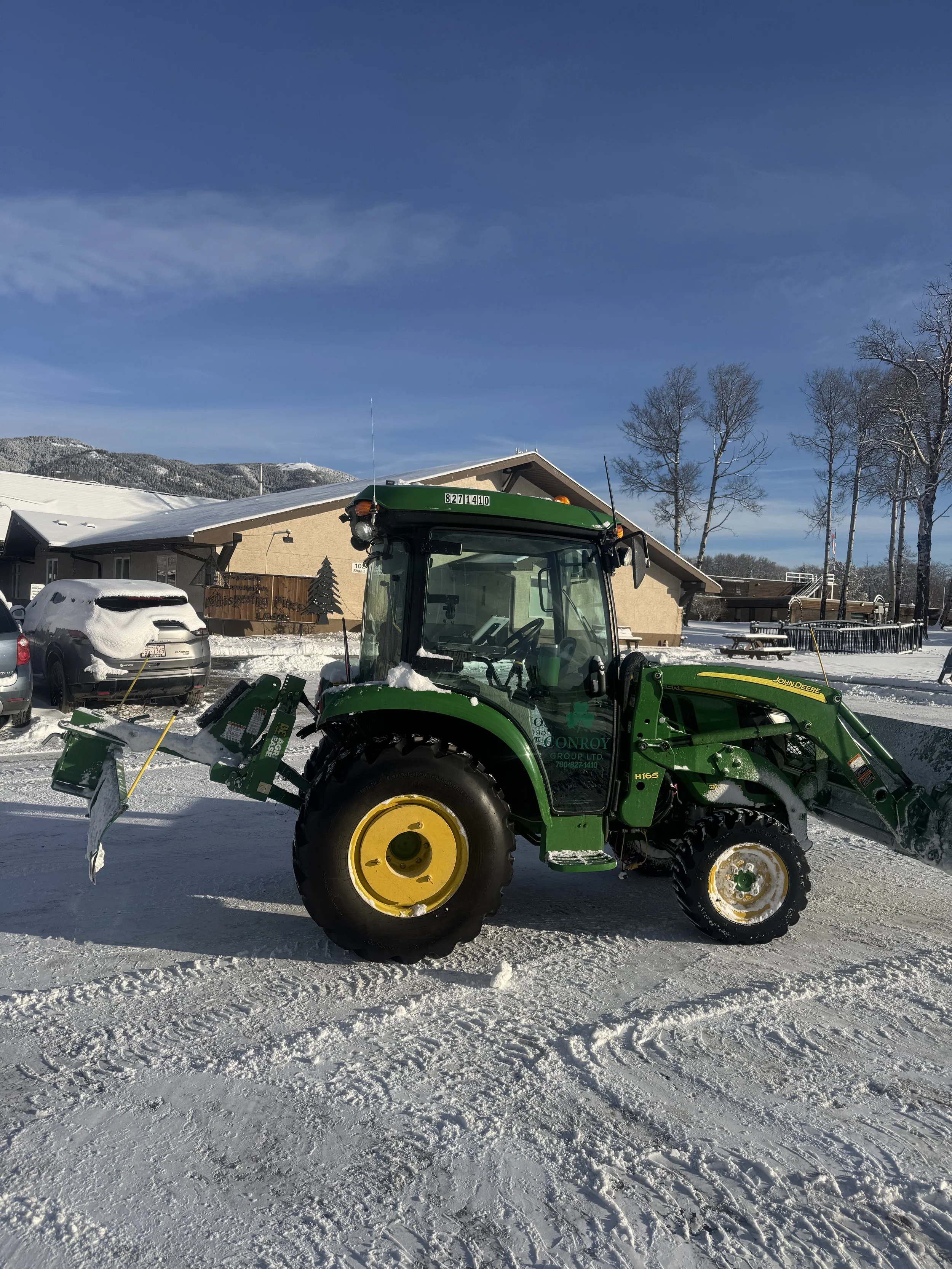 A green John Deere tractor equipped with snow plow attachment parked on snow-covered ground outside a building with snow on its roof, leafless trees, and mountains in the background under a blue sky.