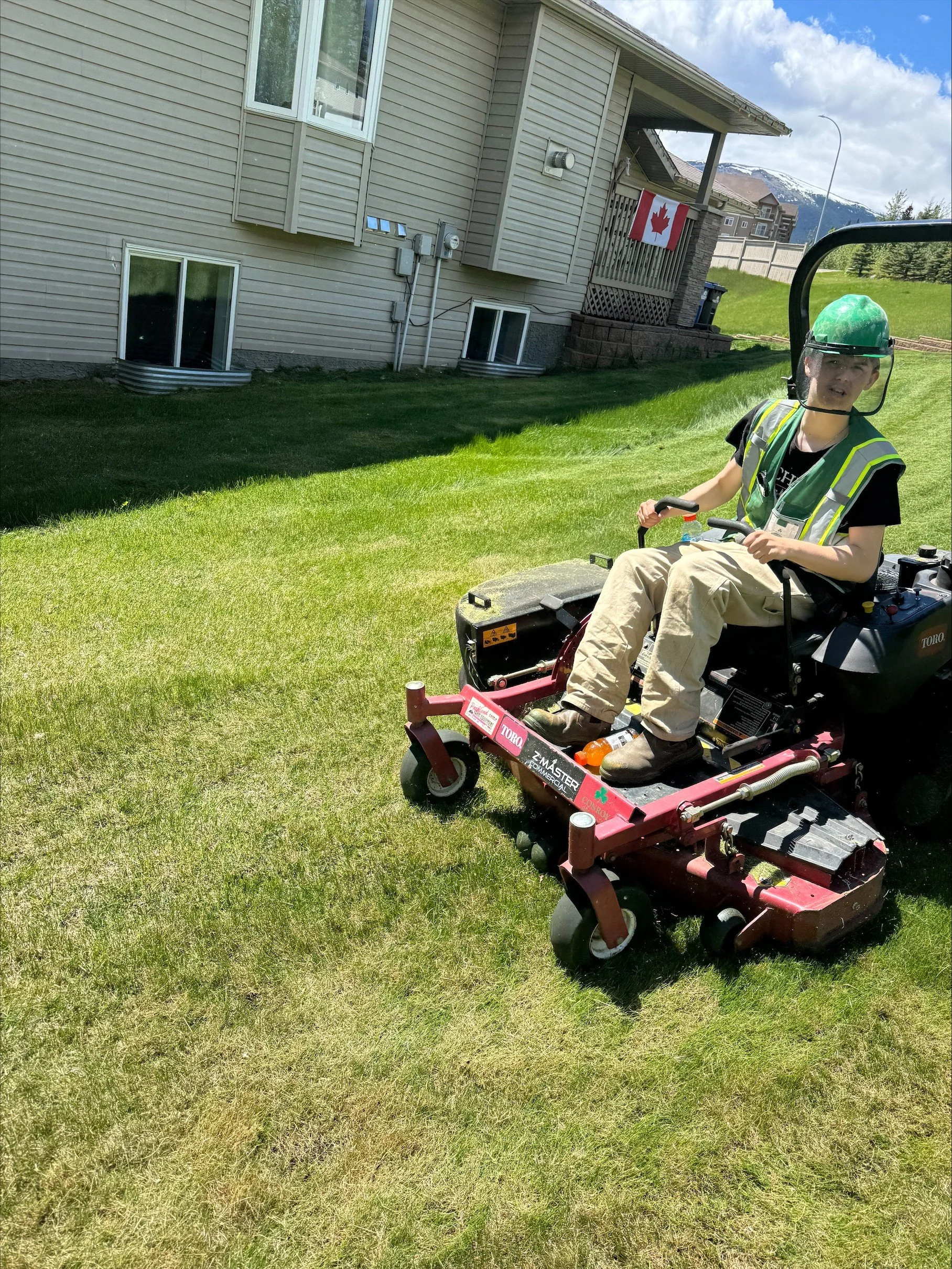 A person sitting on a riding lawn mower in a backyard, wearing a green helmet and safety vest, with a house and mountains in the background.