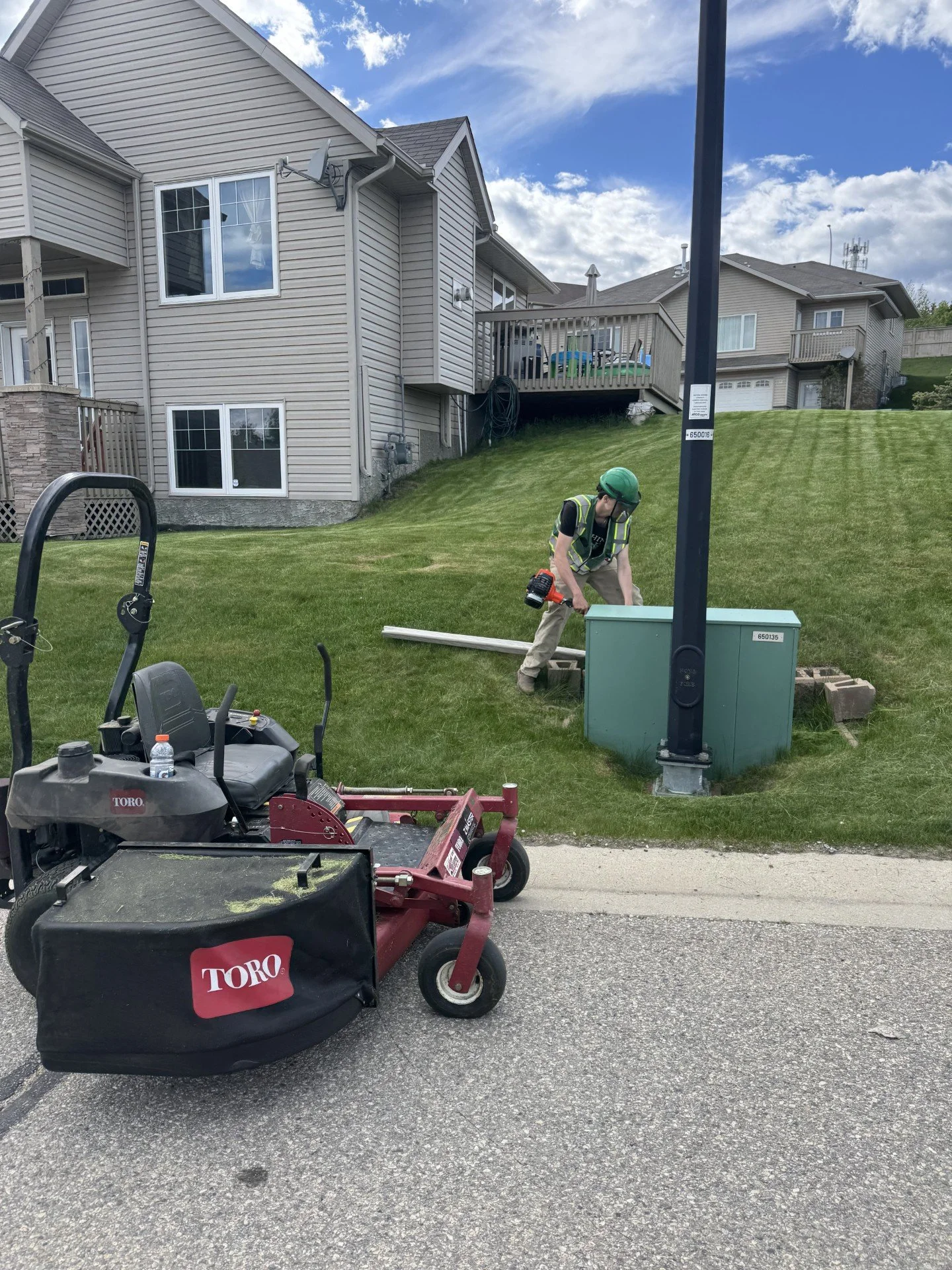 A man wearing a green helmet and safety vest working on an electrical box on a grassy hill in a residential neighborhood. There is a red and black Toro lawn mower in the foreground.