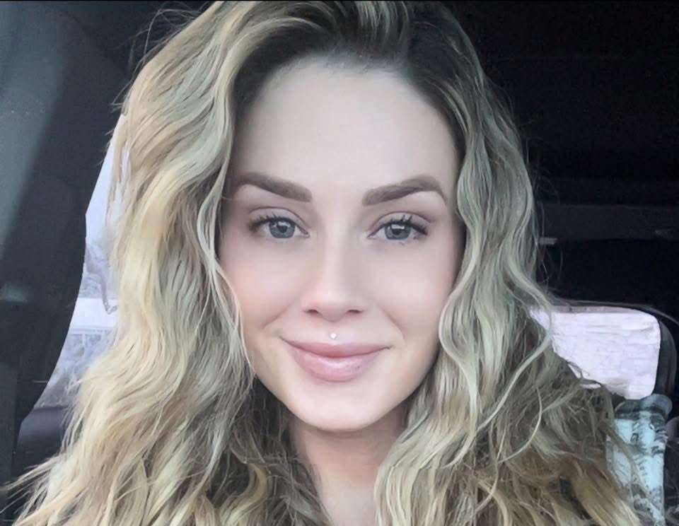 A young woman with wavy blonde hair and blue eyes smiling in a car.