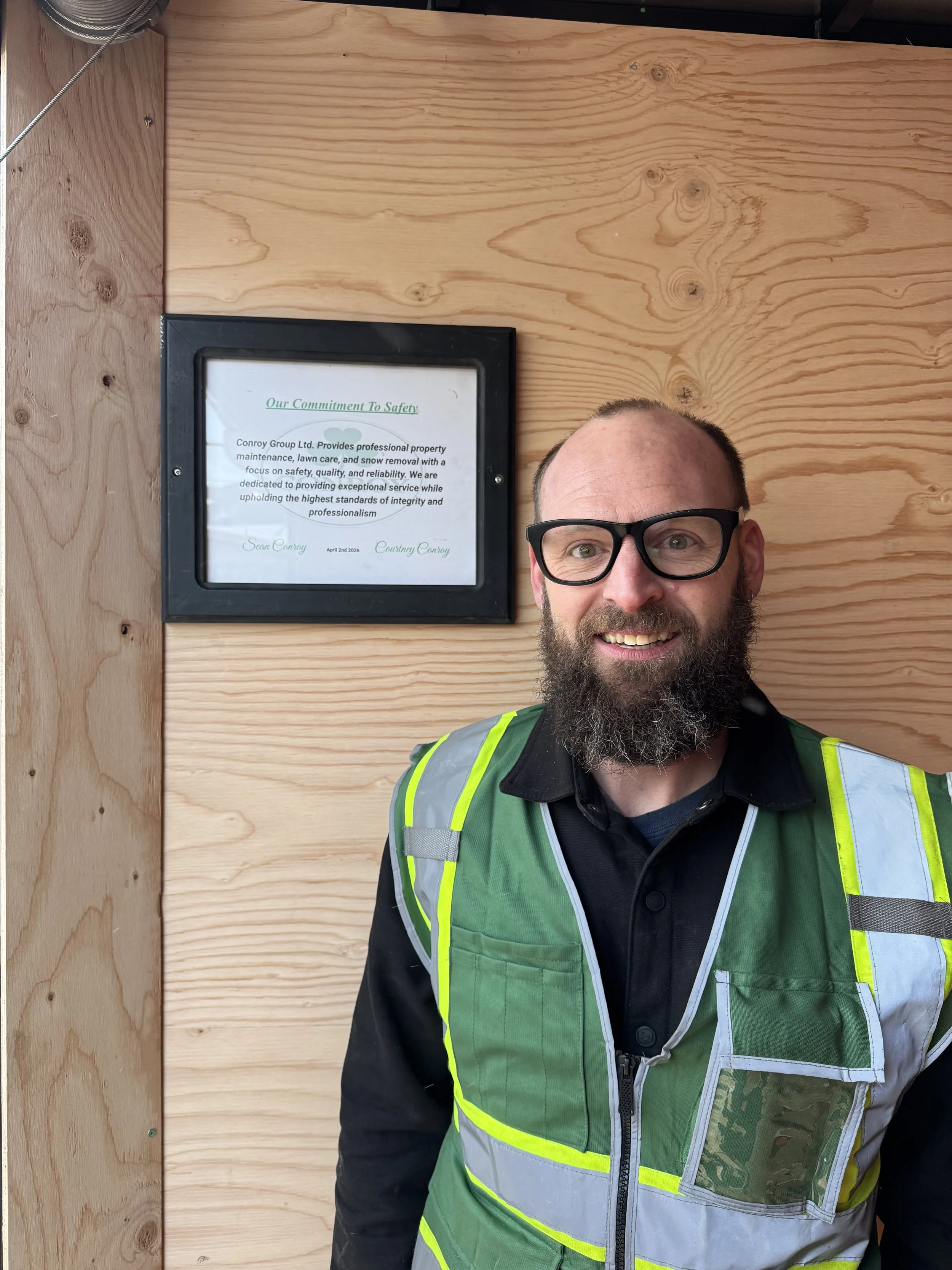 A man with glasses, a beard, and a mustache smiling in front of a wooden wall. He is wearing a black shirt and a high-visibility safety vest. There is a framed document on the wall behind him.