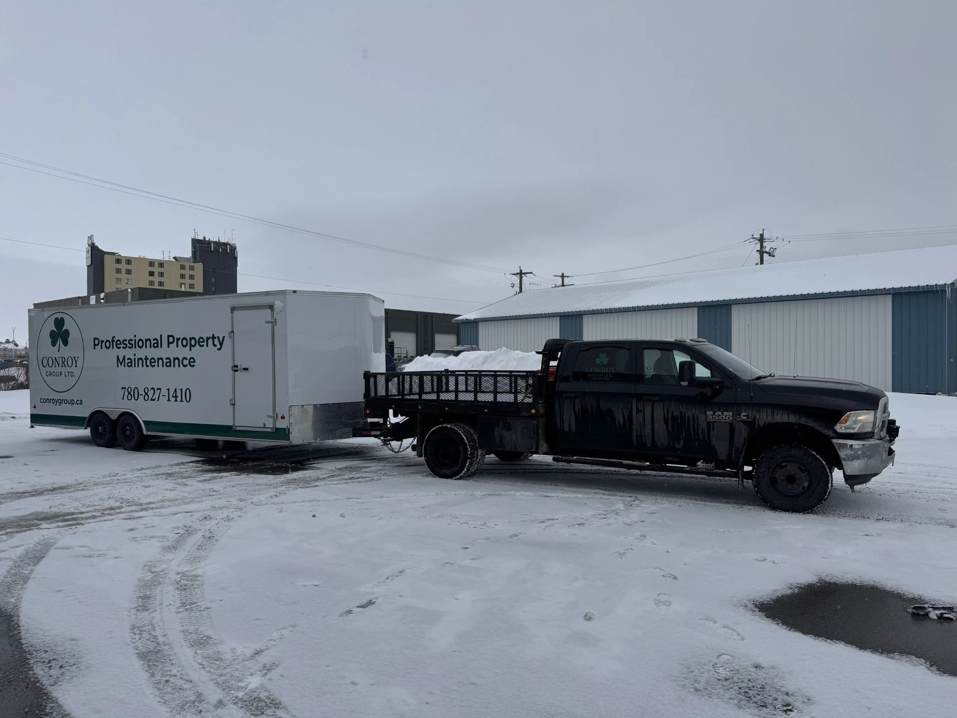 A black pickup truck pulling a white trailer labeled 'Professional Property Maintenance' with contact information, parked on snowy ground near a blue industrial building with a snow-covered roof, and a multi-story building in the background under an overcast sky.