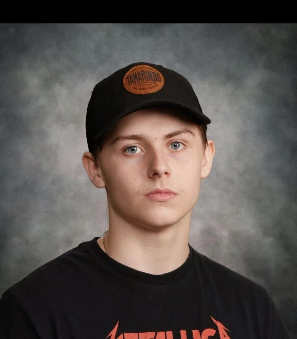 A young man standing outdoors in a backyard with brown pants, a black T-shirt with white 'LA' logo, black cap, and hands in pockets, under a tree with a wooden fence and house in the background.