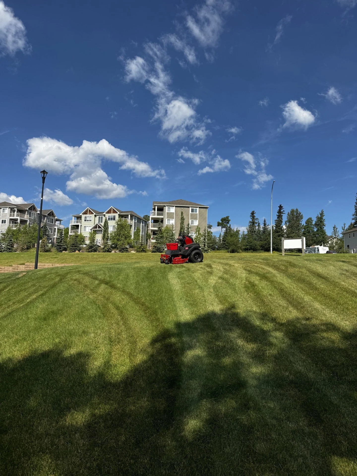 A red lawn mower on well-manicured grass in a residential area with multi-story buildings and trees, under a blue sky with scattered clouds.