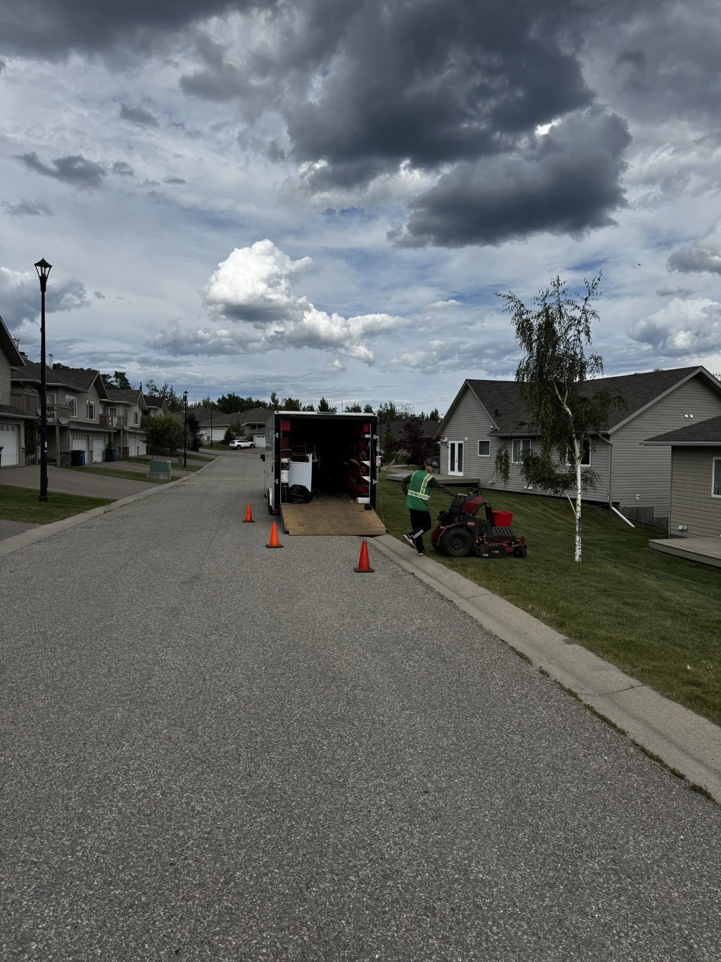 A street with homes on either side, a worker in a green safety vest operating a ride-on lawn mower, and a truck with its back open parked on the street under a cloudy sky.