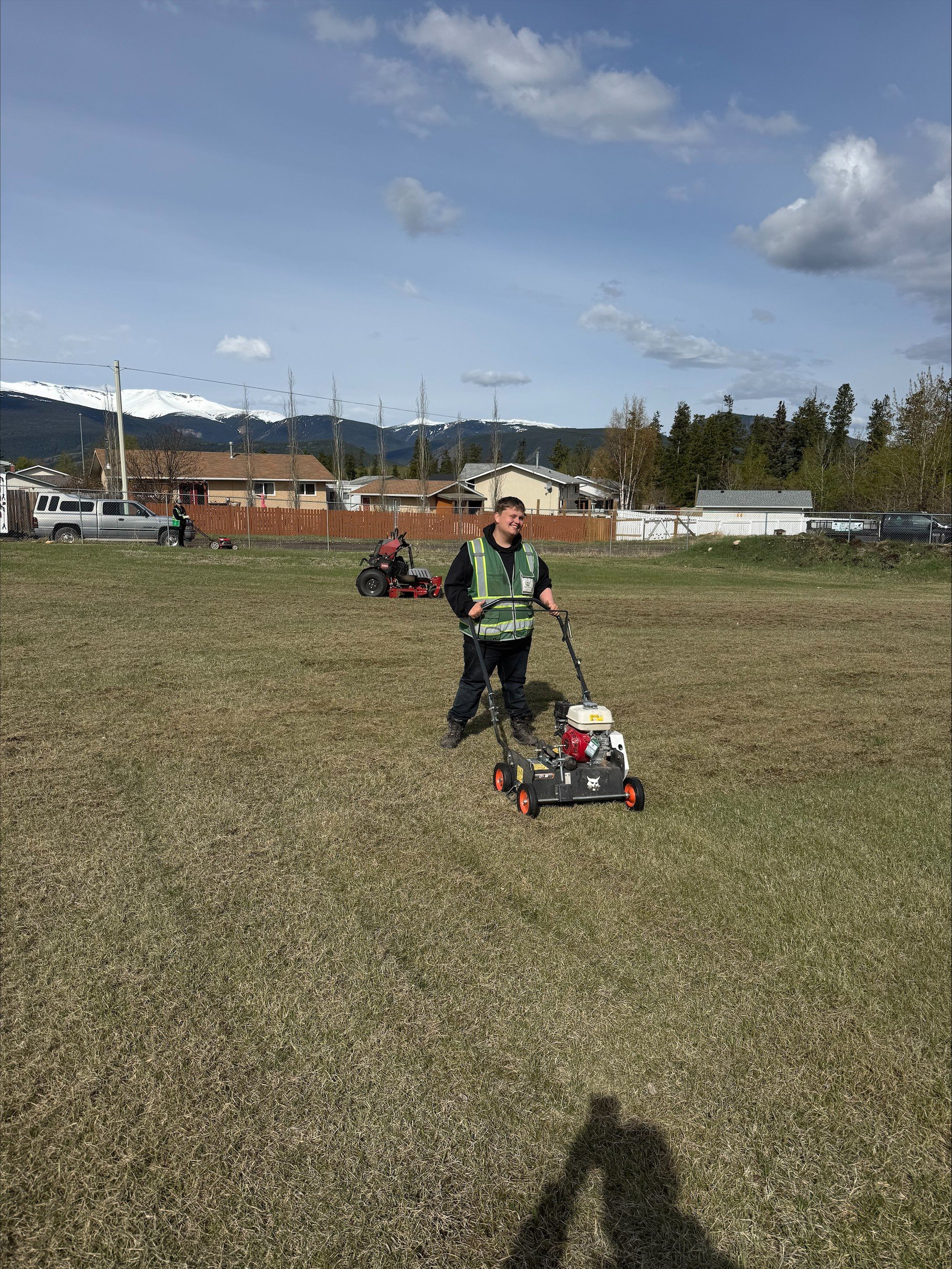 A young man in a reflective vest mowing a grassy field with a push mower. In the background, another person is also working on the field. There are houses, trees, and mountains with snow-capped peaks under a partly cloudy sky.