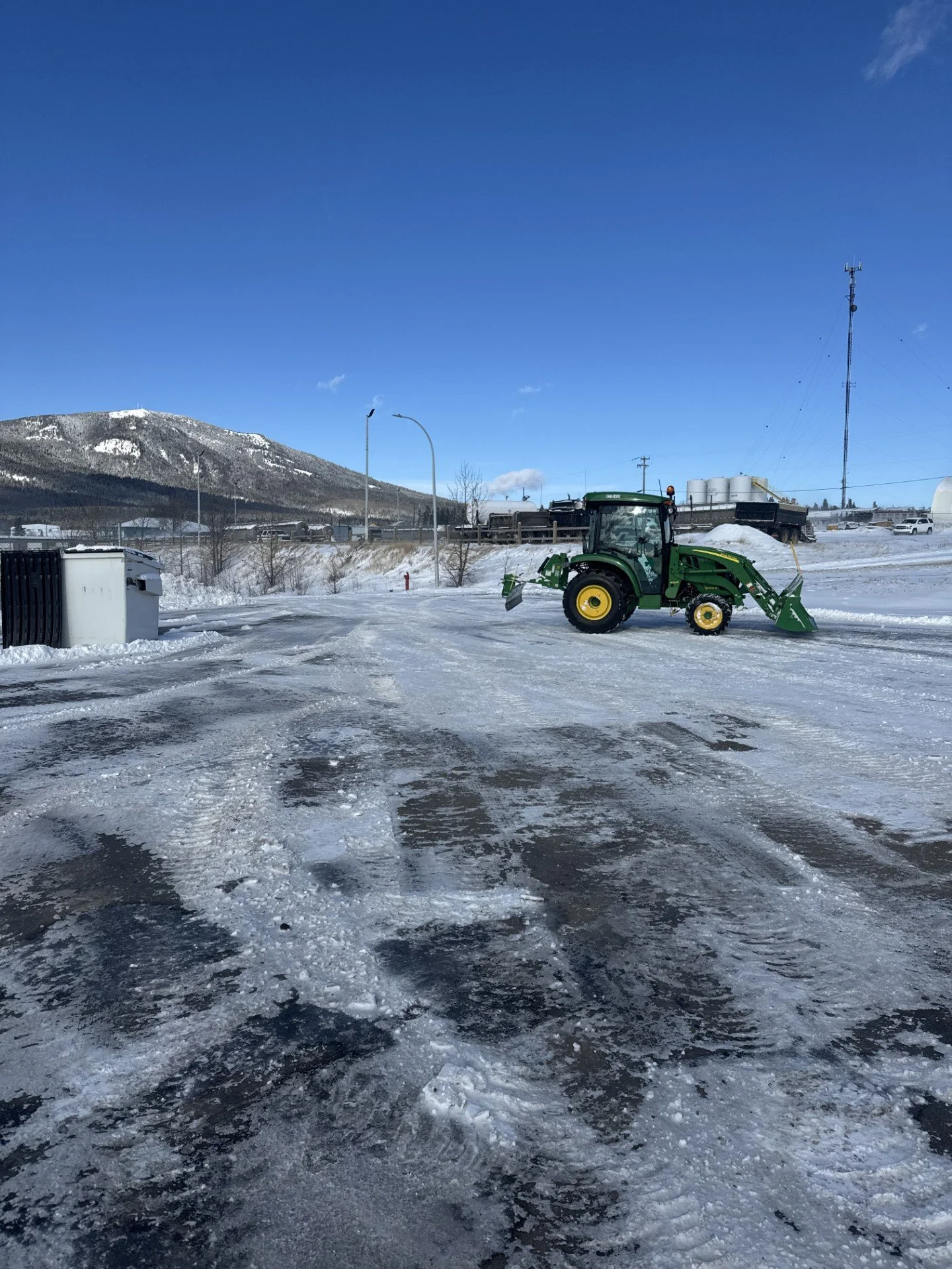 A green tractor with a snow plow attachment clearing snow on a parking lot, with a snow-covered mountain and a clear blue sky in the background.