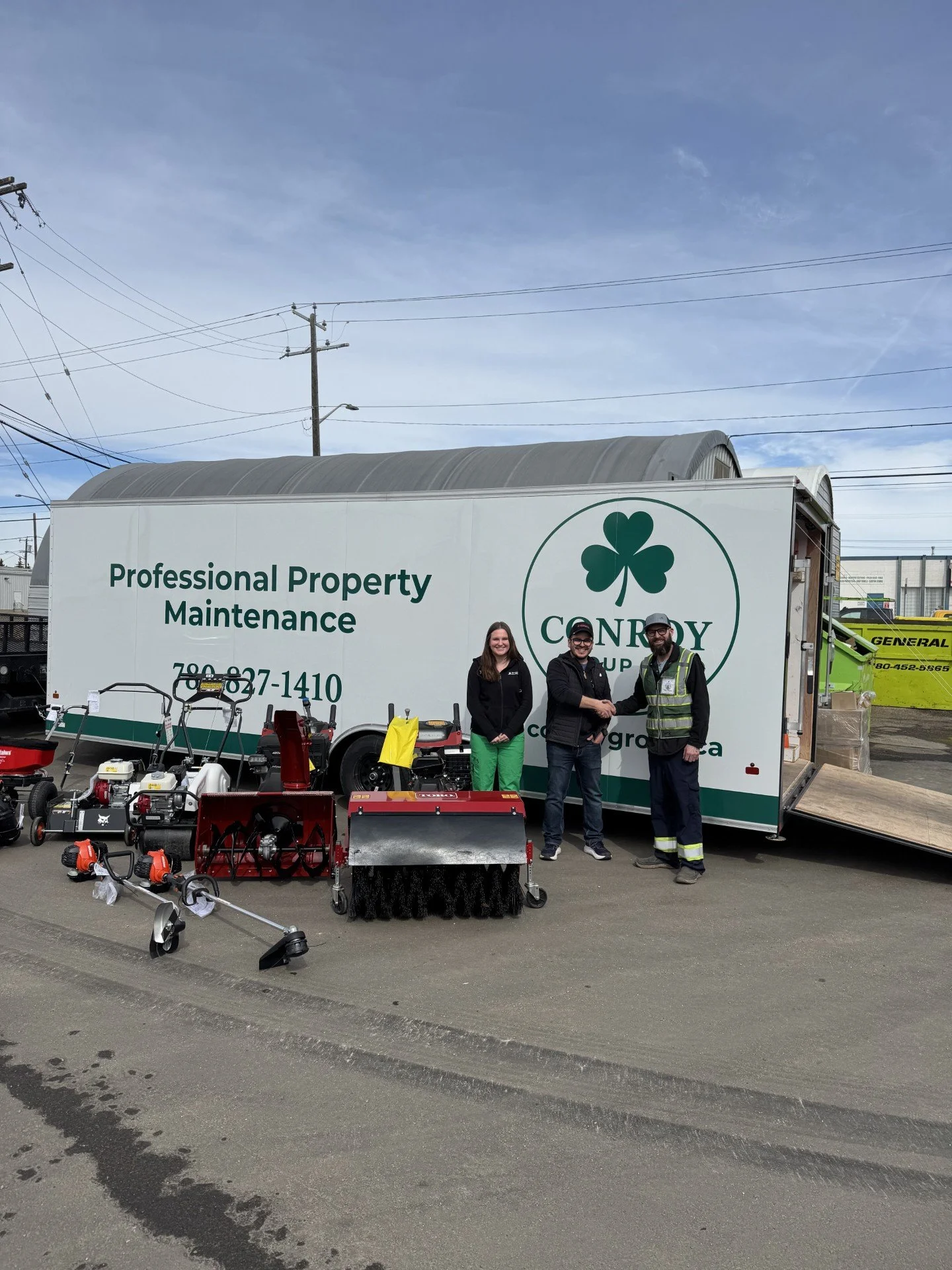 People standing in front of a mobile equipment trailer labeled "Professional Property Maintenance" with various outdoor power tools and equipment displayed, including a gas-powered blower, hedge trimmers, and a snowblower, outdoors on a cloudy day.