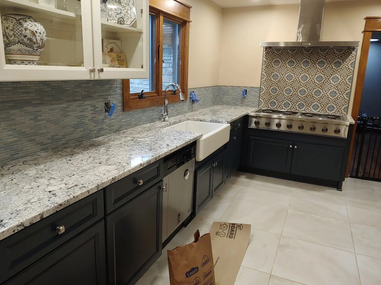 Kitchen with granite countertops, a white farmhouse sink, black lower cabinets, a window above the sink, and a stove with patterned tile backsplash.