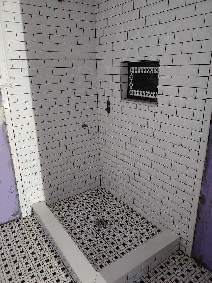 Empty shower stall with white subway tile walls, a small window with black mesh, and black and white mosaic tile floor.