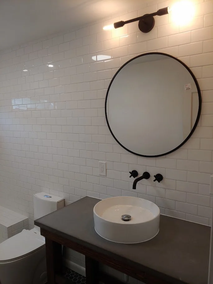 Modern bathroom with white tiled walls, a round mirror above a black faucet, a vessel sink on a wooden vanity, and a toilet to the side.