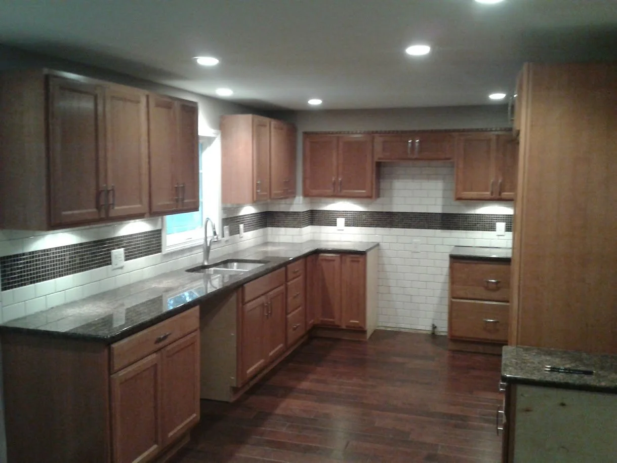 Kitchen with wooden cabinets, granite countertops, a window over the sink, and tile backsplash, illuminated by recessed ceiling lights on dark hardwood floor.