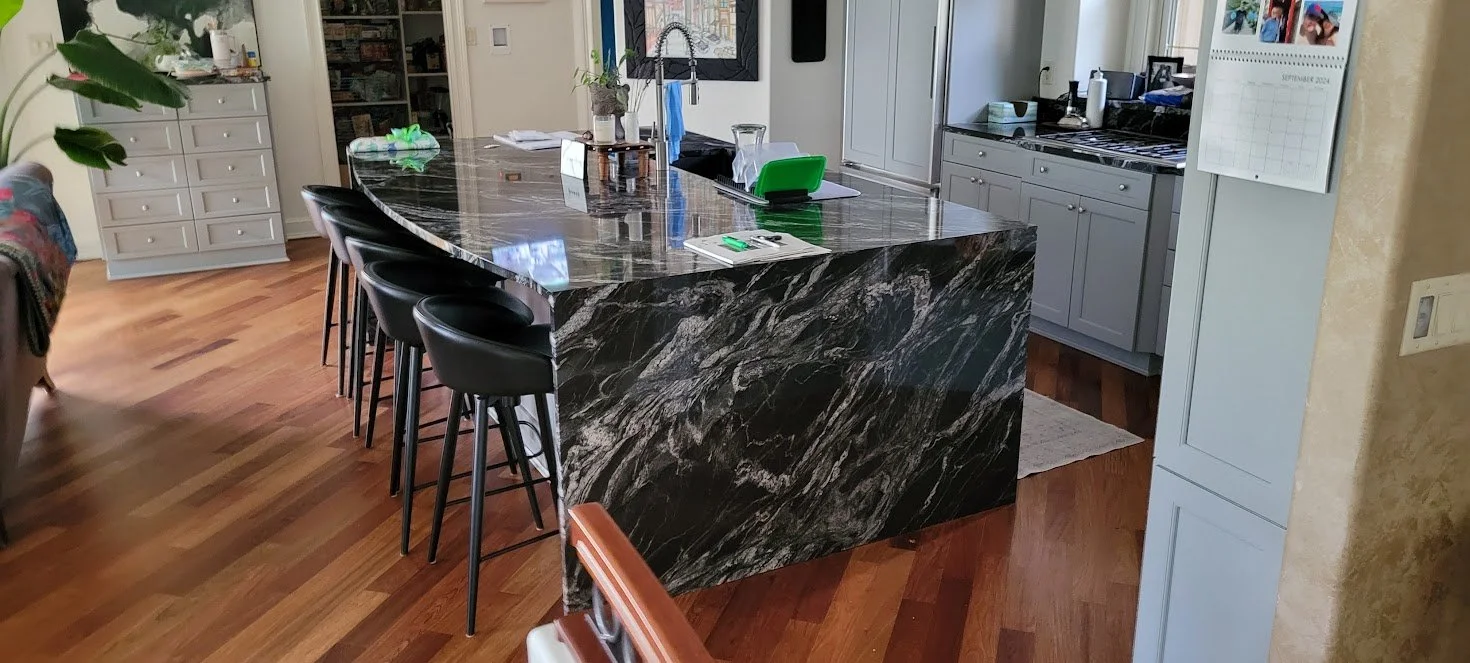 Modern kitchen with a large black marble island, black bar stools, gray cabinets, and hardwood floors.