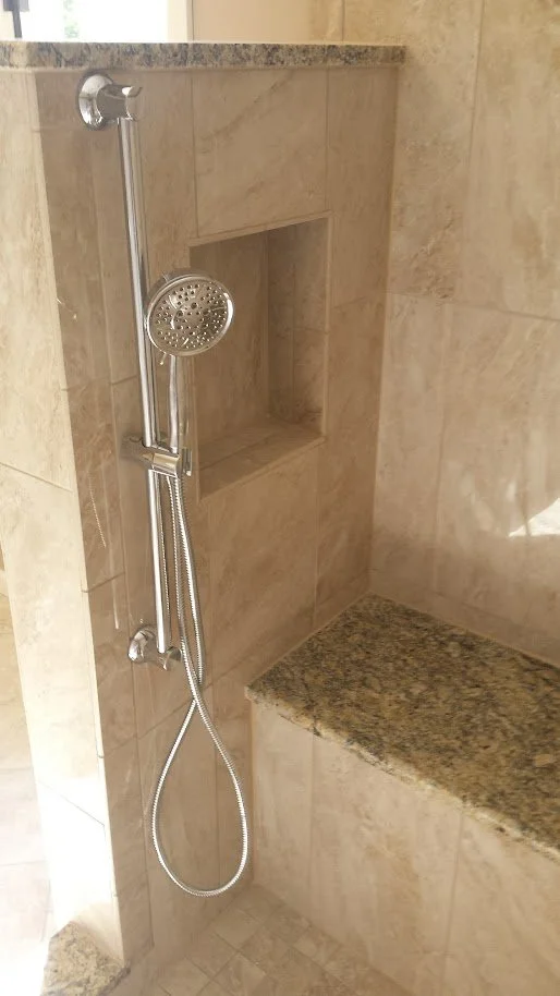 A shower area with beige tiled walls, a built-in niche, a chrome showerhead with a handheld attachment, and a granite bench.