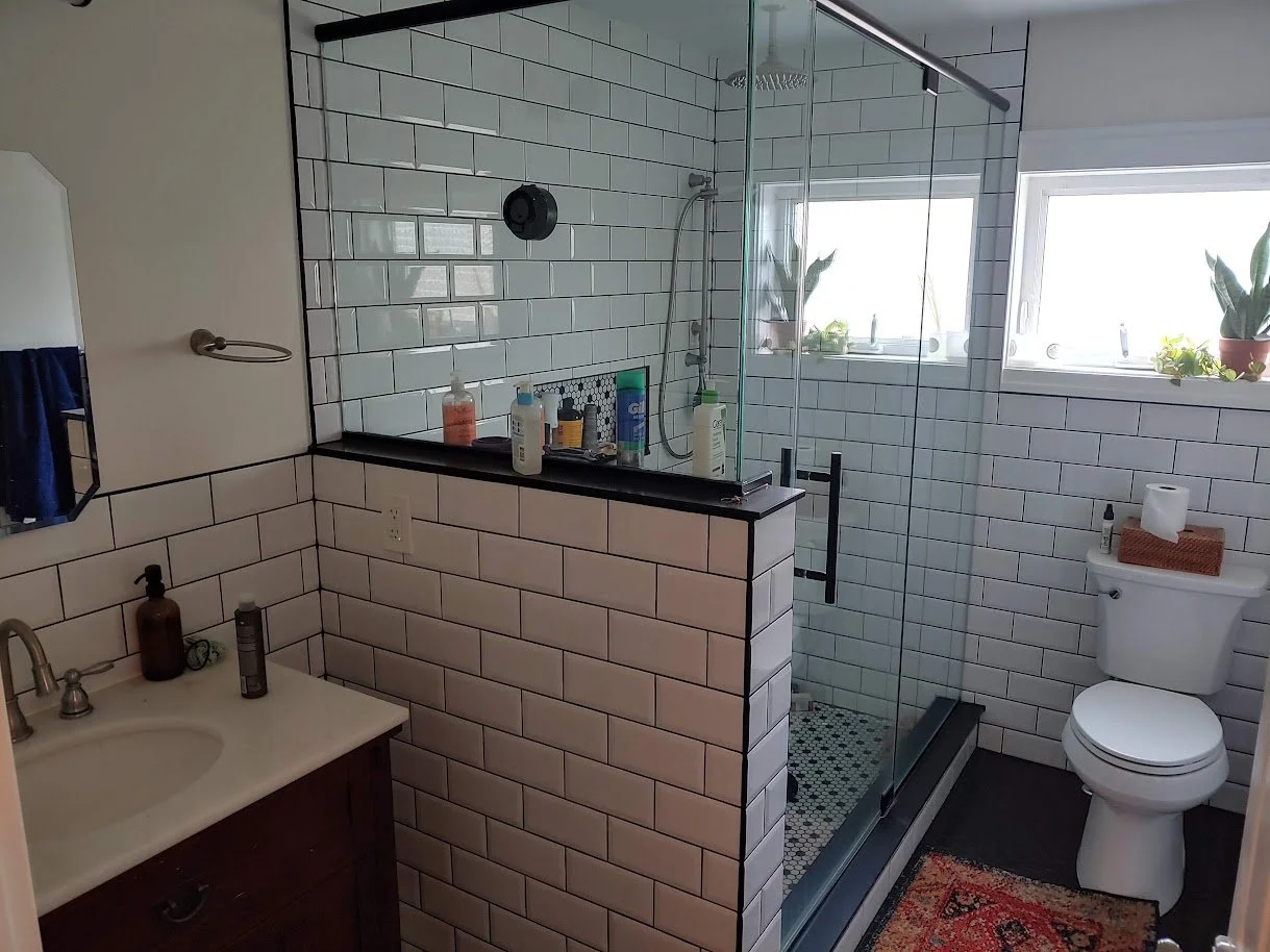 Bathroom with white subway tile walls, glass shower with black hardware, and a toilet with a wicker tissue box on top. There are two windows with potted plants, a small sink, and personal care products on a black-topped half-wall.