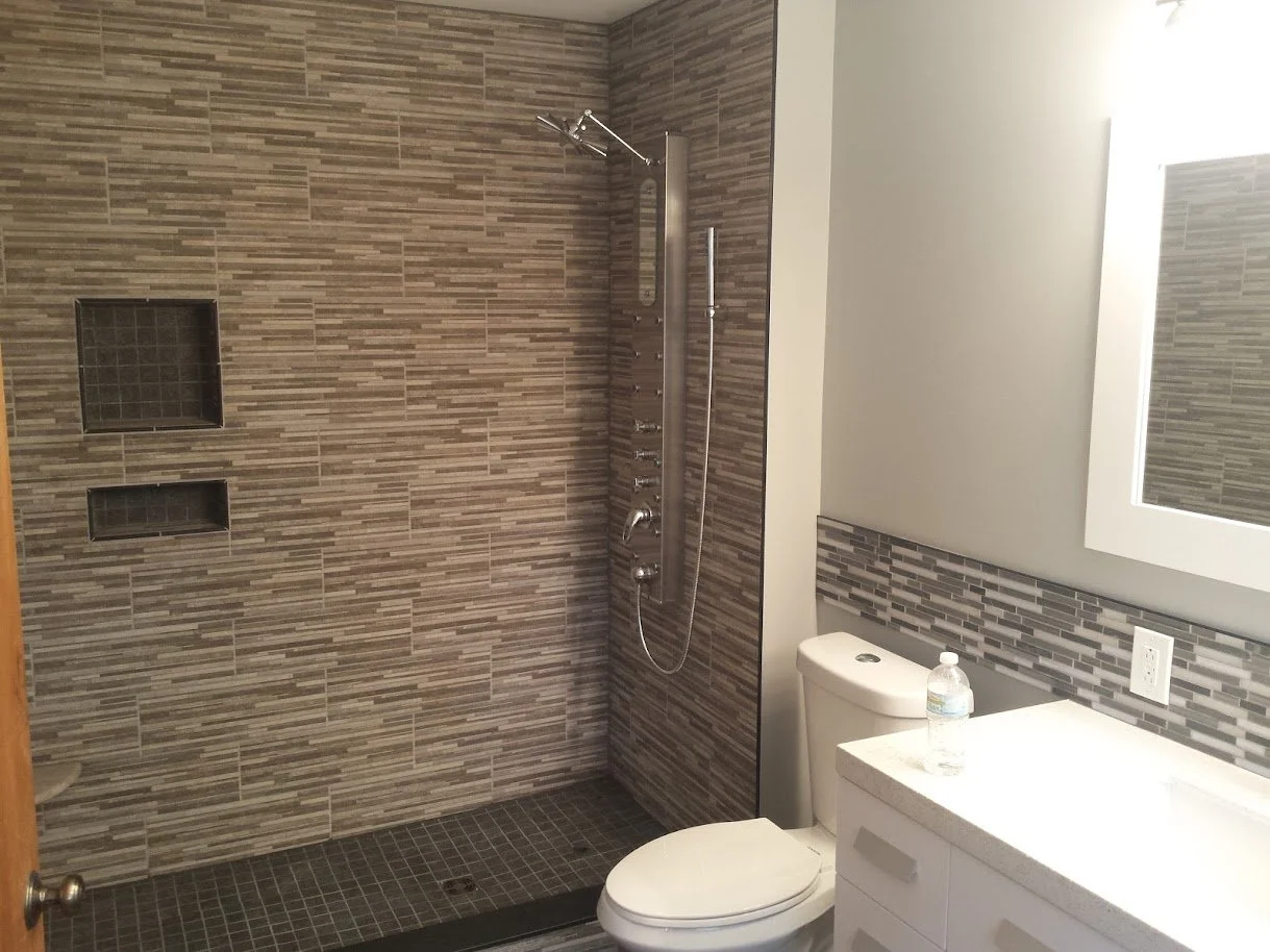 Bathroom with a walk-in shower, toilet, and mirror with a vanity. The shower has two built-in niches, a rainfall showerhead, and a handheld shower attachment, with brown and gray tiled walls and dark tiled floor.