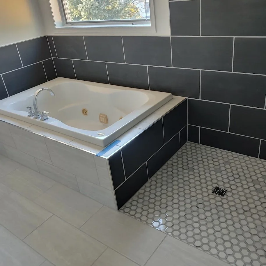 A white bathtub with chrome faucet and jets, surrounded by black tile walls, next to a window, with a separate small hexagonal tiled shower area with a drain on the floor.