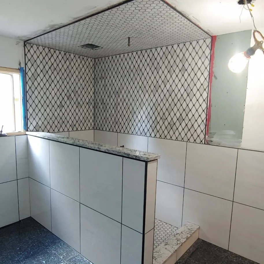 Interior of a bathroom under renovation with white wall tiles, a partial granite counter, a small window, and a paper-covered section of wall with a light bulb overhead.