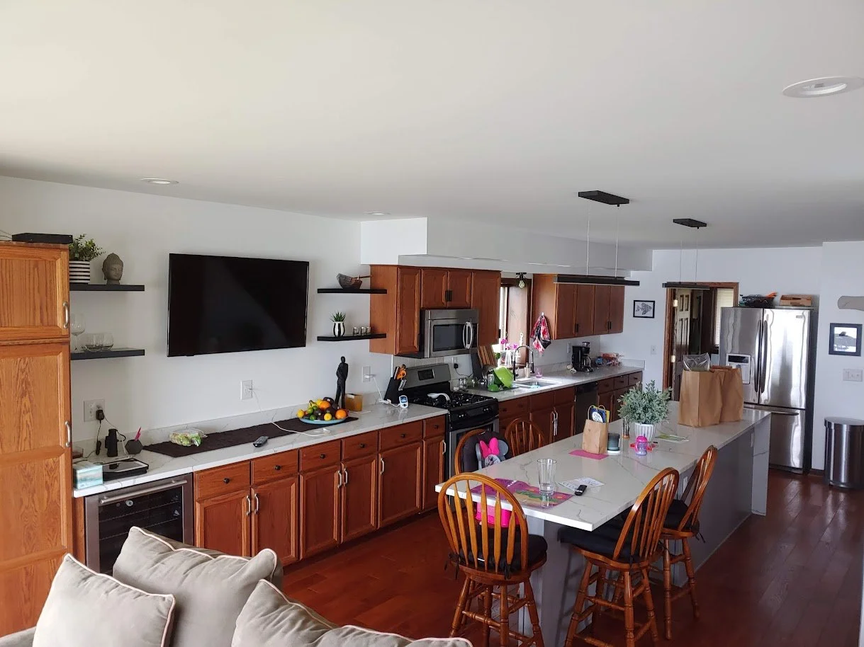 Open kitchen with wooden cabinets, stainless steel appliances, a white island with chairs, a wall-mounted TV, and decorative shelves with plants and art.