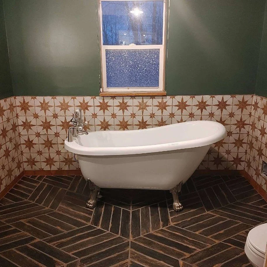 Bathroom with a white vintage clawfoot bathtub, dark patterned tile floor, decorative tile wall with starburst design, green walls, and a window with raindrops on the glass.