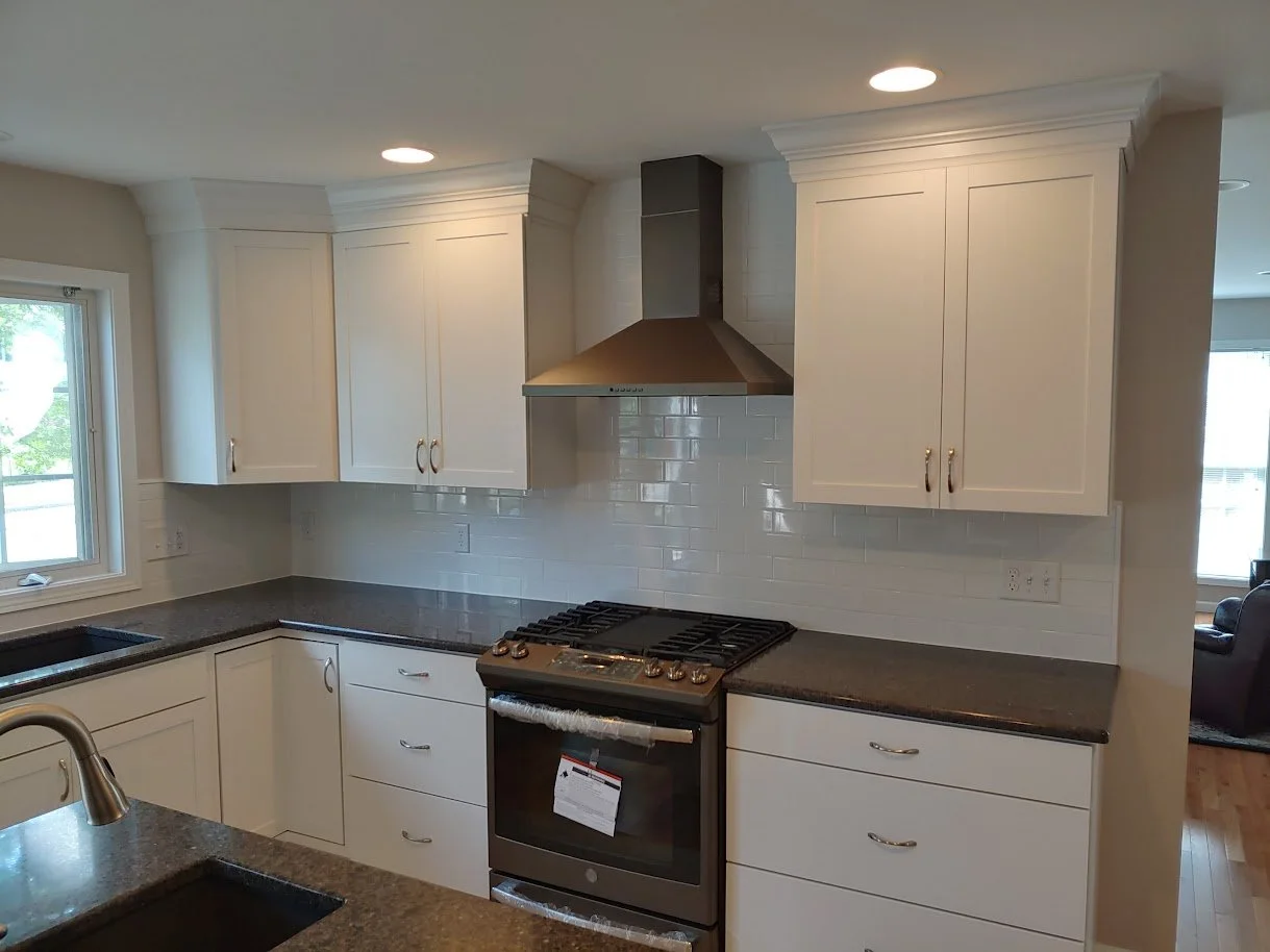 Modern kitchen with white cabinets, black countertops, stainless steel stovetop and range hood, white backsplash, and a window overlooking the outdoors.