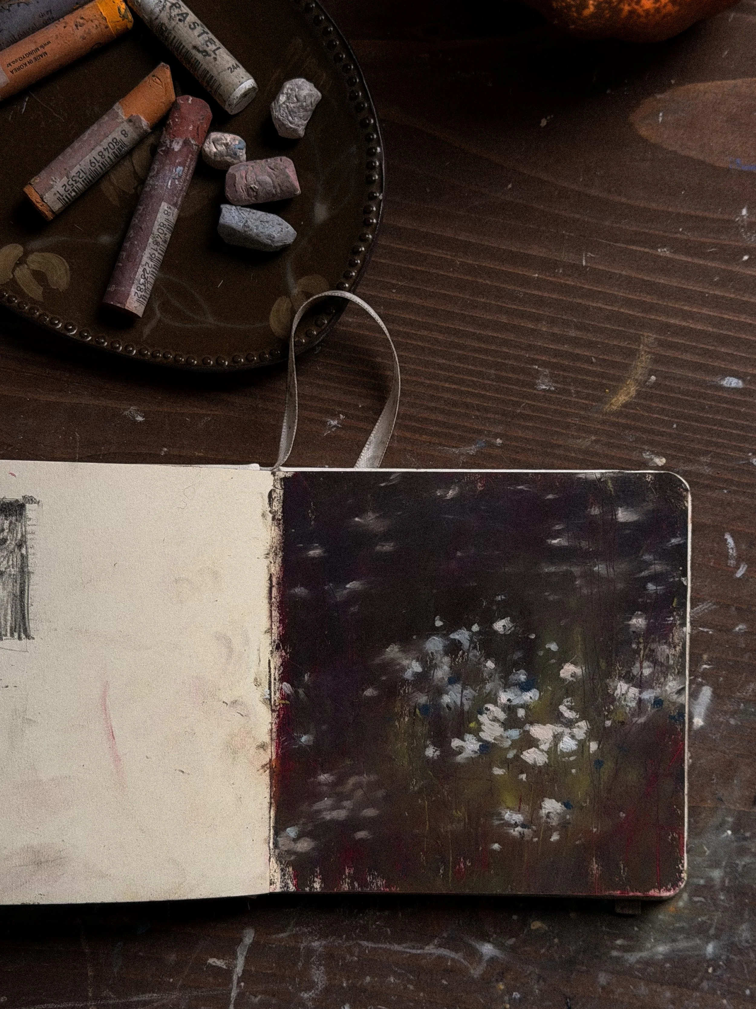 A sketchbook open to a page divided into four sections, with a color chart, a white background, and a black painted area. A dark wooden surface is underneath the sketchbook, and there is a palette with dried paint and some chalk pastels or paint sticks nearby.