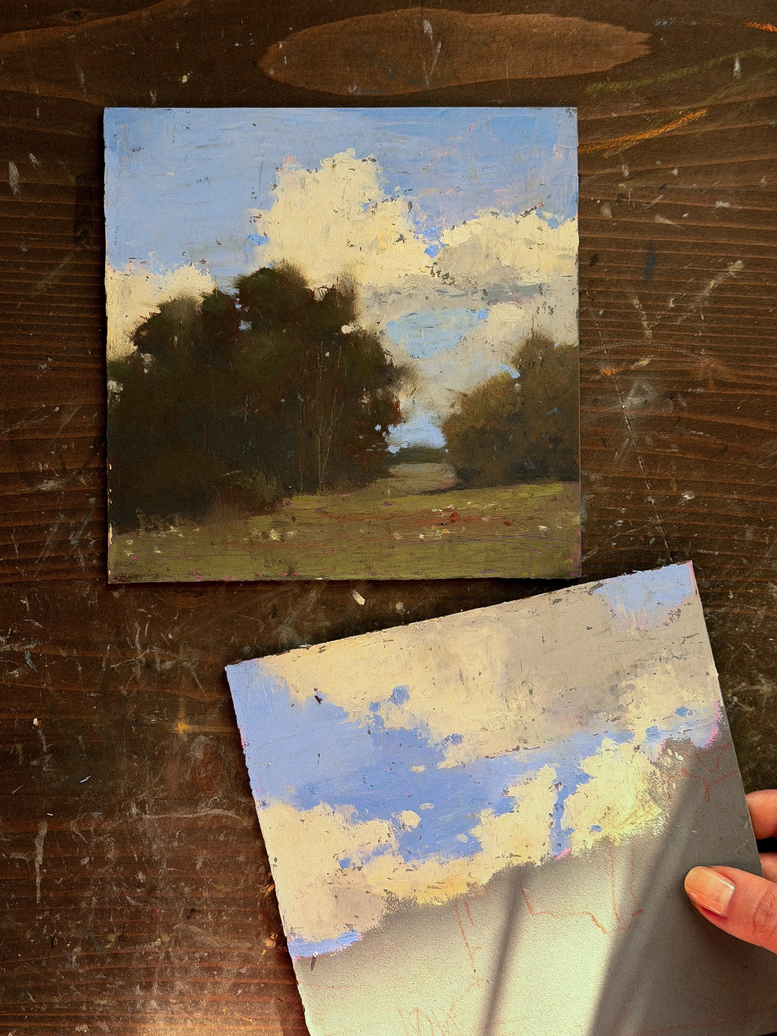 Landscape painting of trees, sky, and clouds, with brushstrokes visible, on a wooden surface.