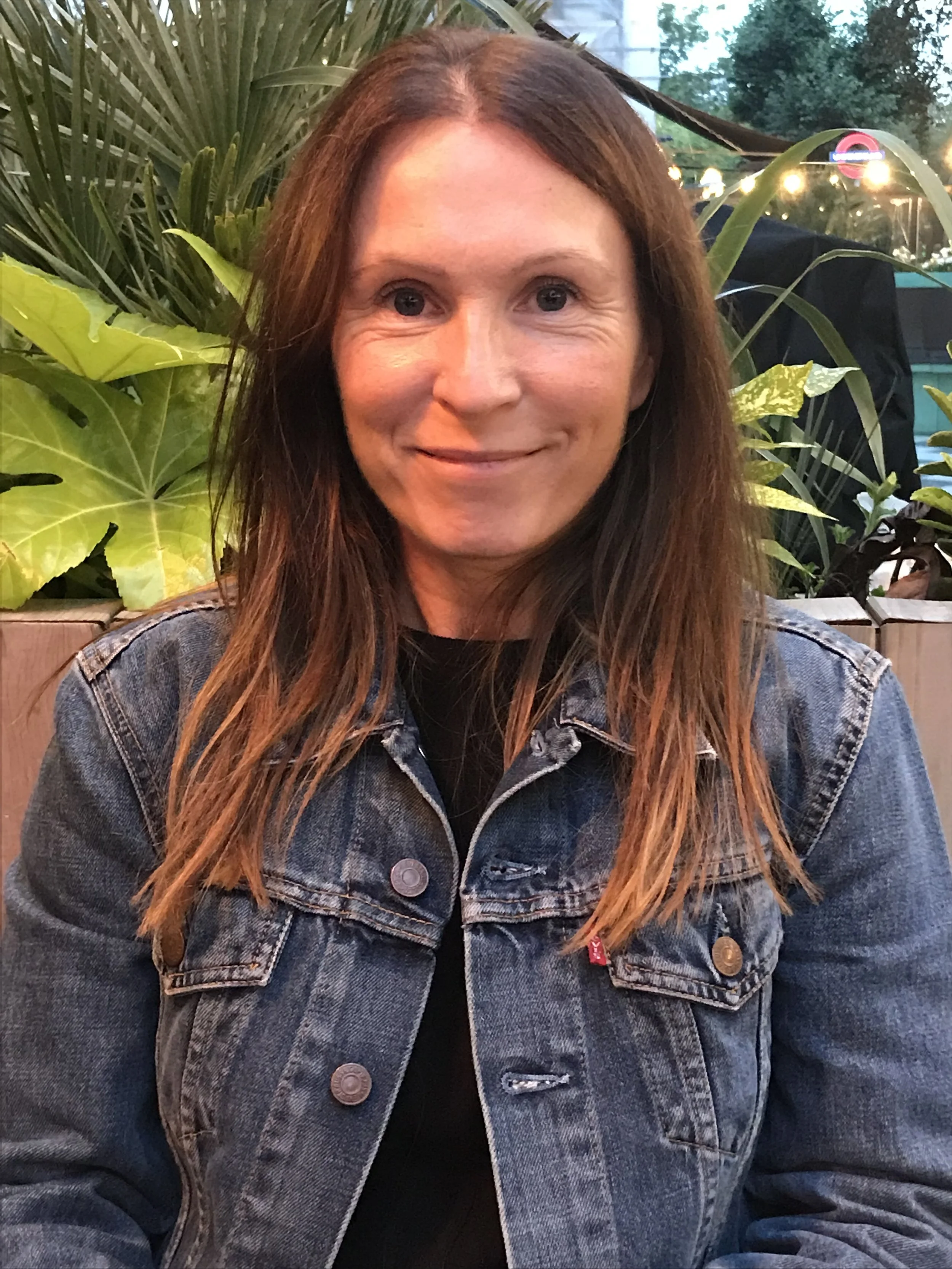 A woman with long brown hair and a denim jacket sitting outdoors at a restaurant or cafe, greenery in the background, smiling at the camera.