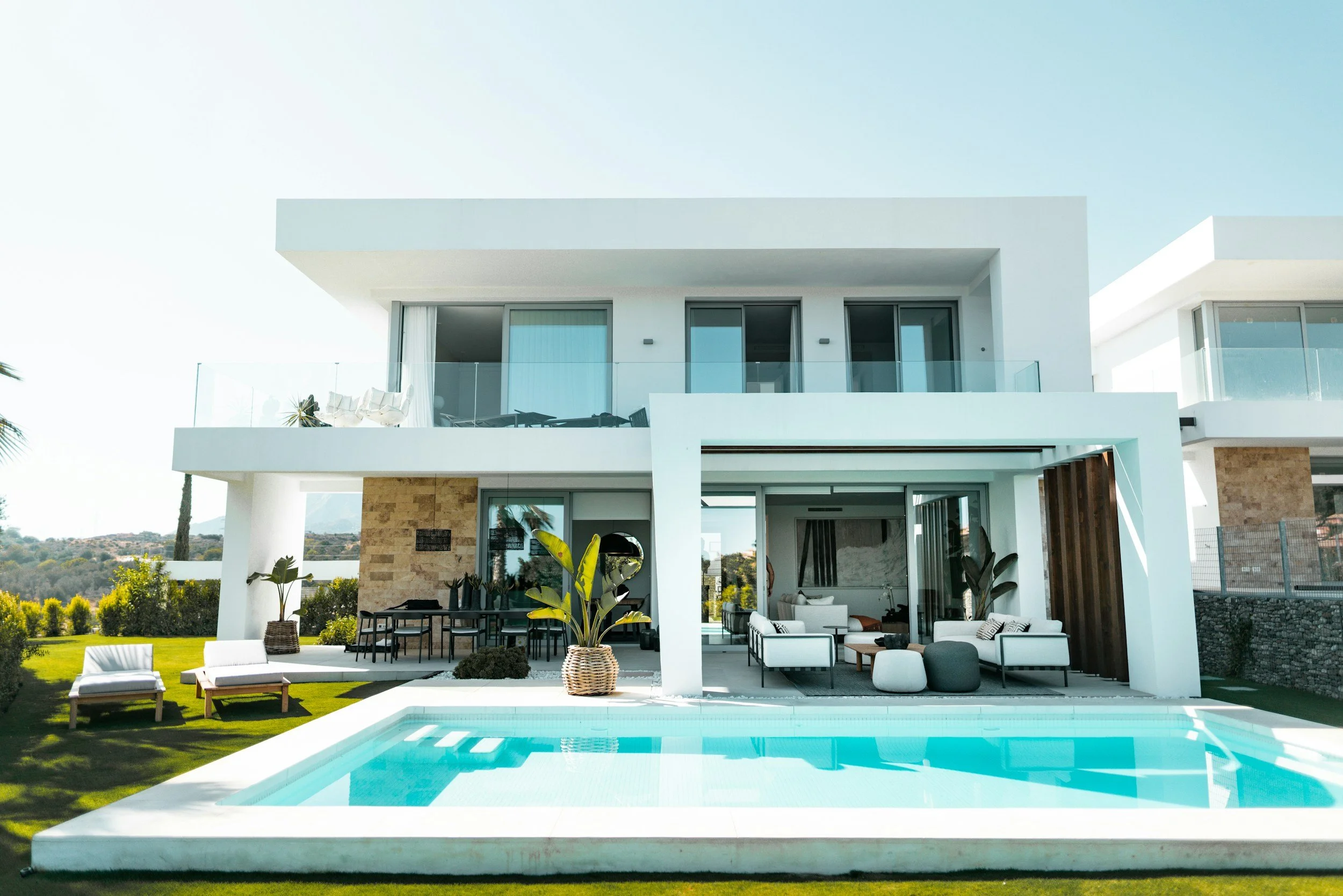 Modern white house with large windows, an outdoor patio area with furniture, and a swimming pool in the foreground, surrounded by green lawn and landscape.