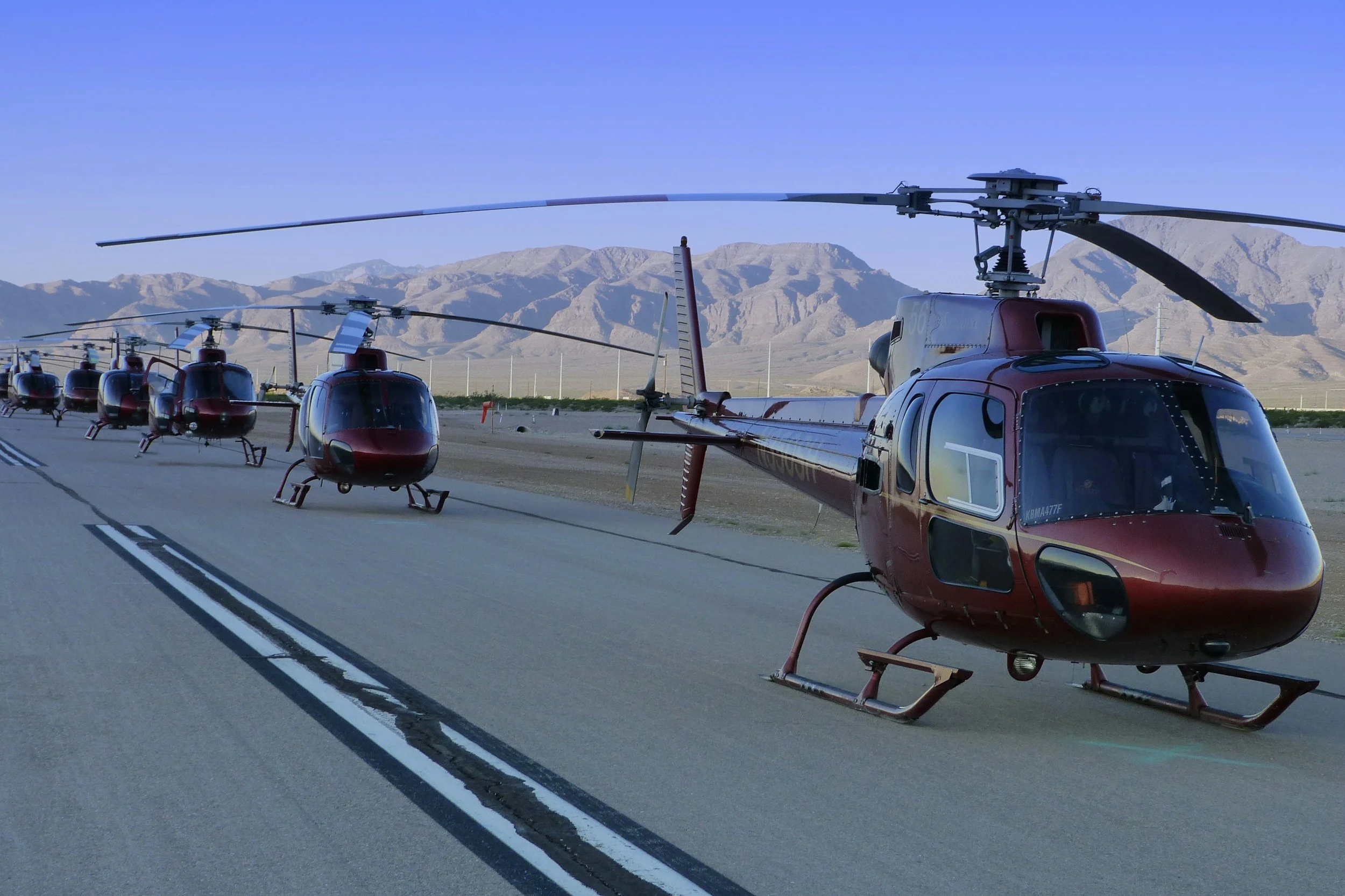 Several red helicopters parked in a row on a runway with mountains in the background under a clear blue sky.