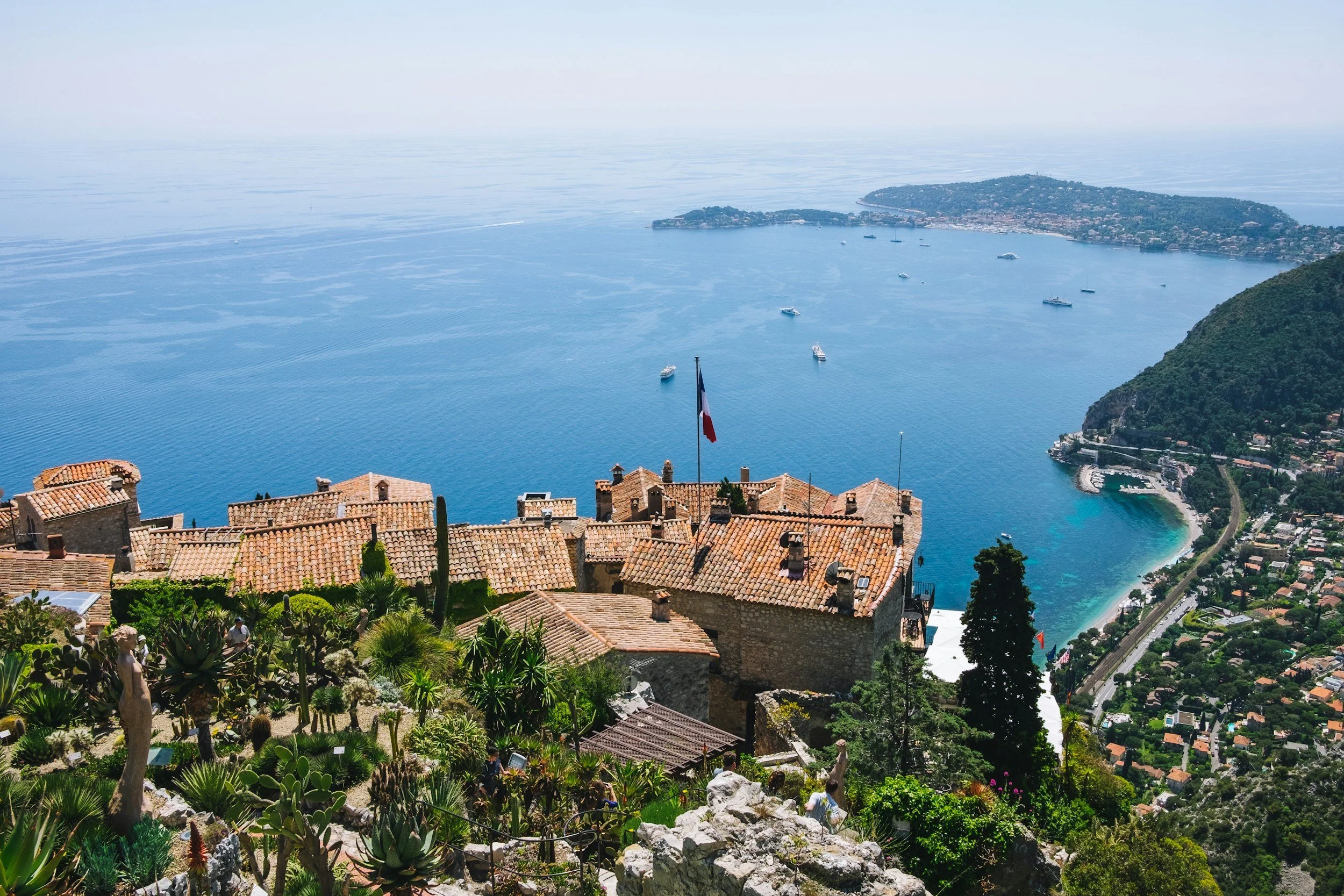 A scenic view of a coastal town with terracotta rooftops, lush greenery, and the ocean in the background. Several boats are anchored in the water, and a flag is visible in the foreground.