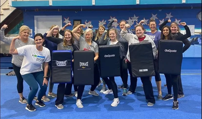 Group of women in an exercise or martial arts class demonstrating strength by flexing their biceps while holding padded shields, in a gym with blue mats and decorative stars on the wall in the background.