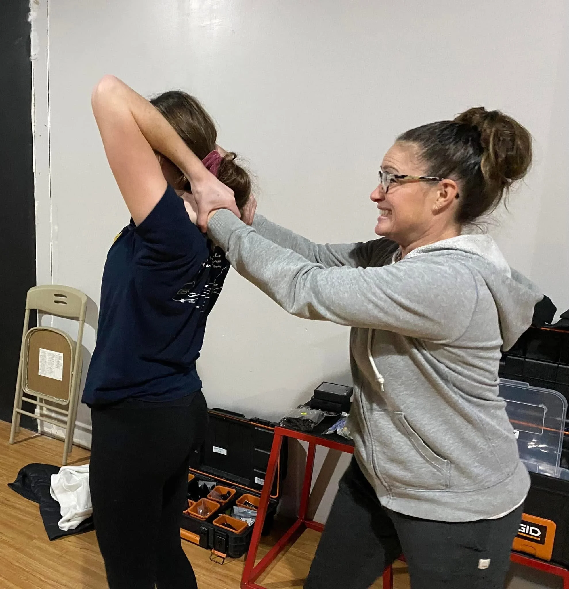 Two women engaged in a self-defense training exercise inside a room. One woman is grabbing the other by the face while the second woman is reacting defensively.