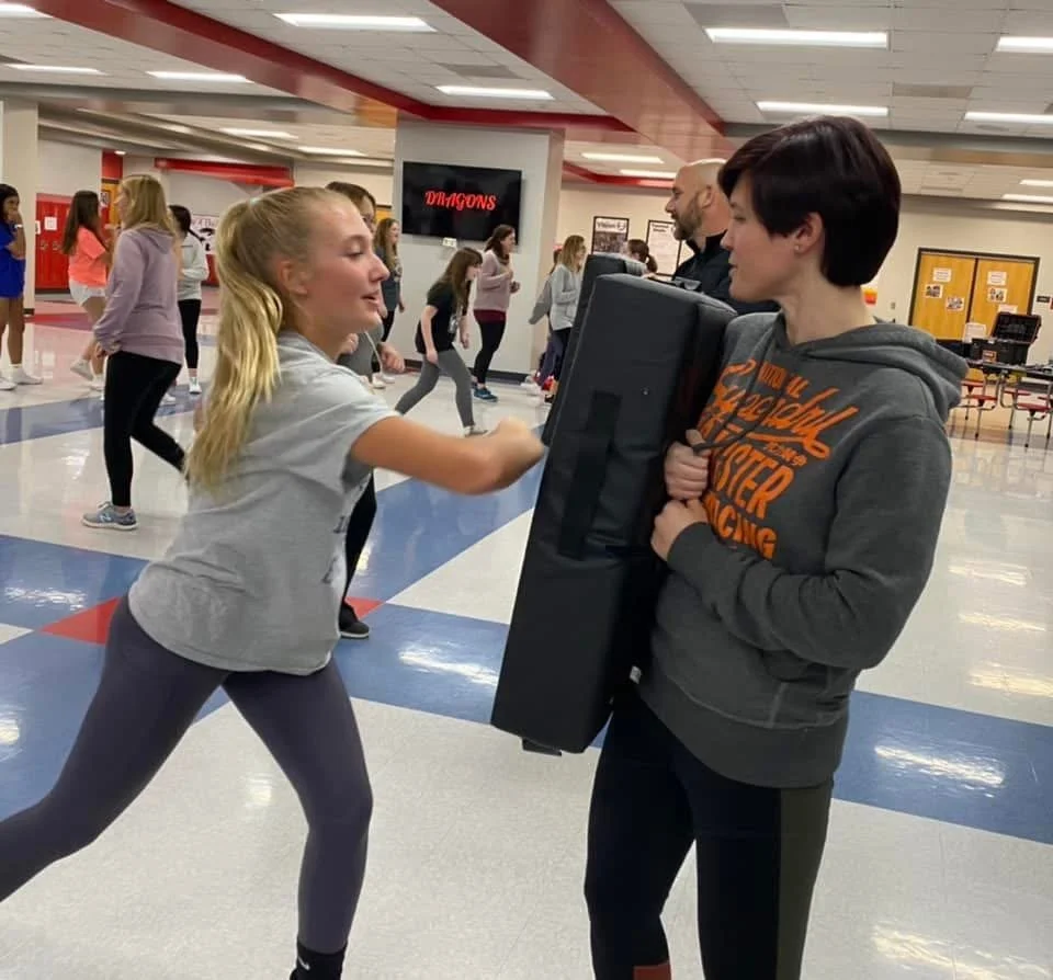 A girl practicing self-defense with a woman holding a padded shield in a school hallway.