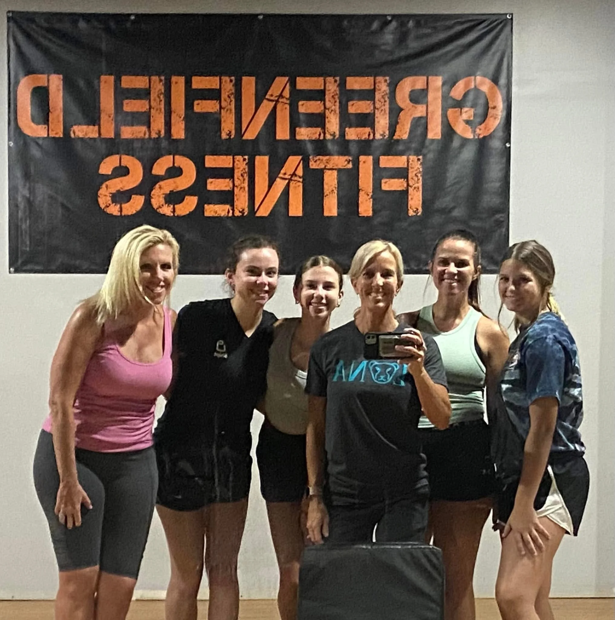 Group of six women in workout attire standing in front of a black and orange sign that reads "GREENFIELD FITNESS" with some women smiling at the camera.