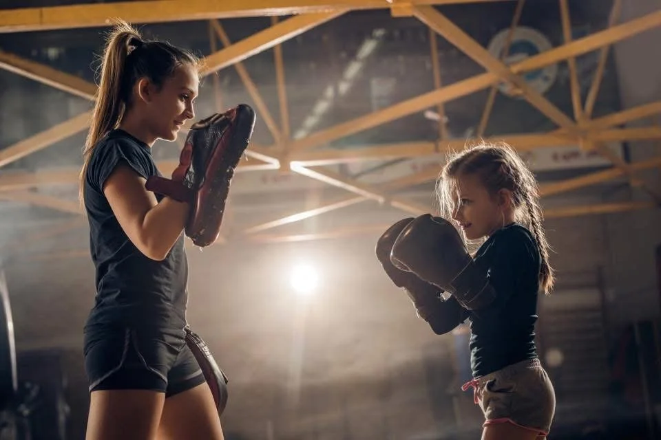 Two young girls practicing boxing in a gym, facing each other with boxing gloves. The gym has a high ceiling with wooden beams and bright lighting.