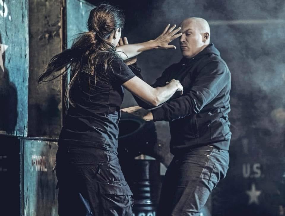 A woman and a man practicing self-defense techniques in a training gym. The woman is blocking the man's punch while he tries to restrain her.