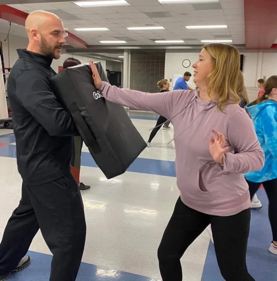 A woman throws a punch at a side-looking punching pad held by a man in a gym or fitness class setting.
