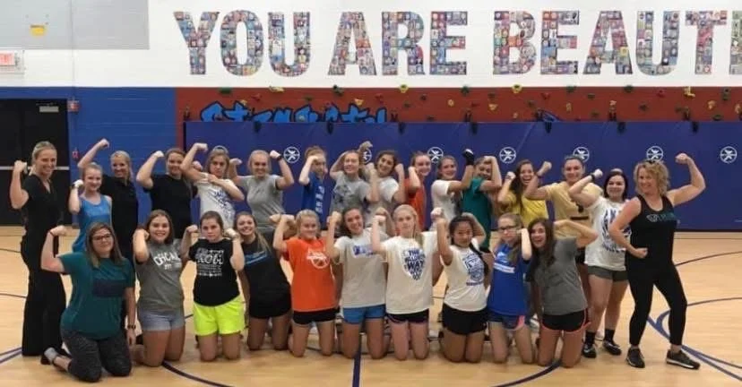 Group of young girls and women in a gymnasium posing with flexed arm muscles, with a large colorful sign that reads 'You Are Beautiful' in the background.